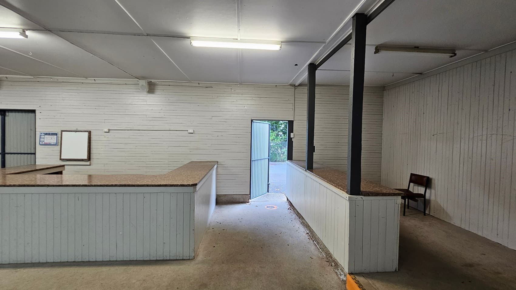 A room with light green wooden walls and a white ceiling. On the left, there is a counter with a speckled brown surface. A notice board and a closed door are visible on the left wall. In the centre, an open door leads outside, showing greenery. On the right, a raised platform with a similar counter and a single red chair. The floor is concrete, and fluorescent lights are mounted on the ceiling.