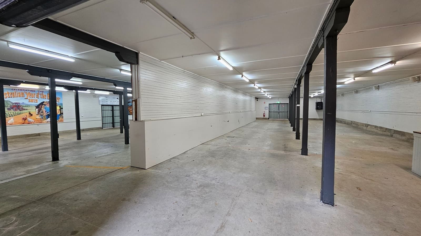A spacious, empty indoor area with a concrete floor and a white panelled ceiling. On the left, there are black metal support beams and a mural with agricultural imagery and text. The right side features a long white wall. At the far end, a metal gate and a fire extinguisher are visible. The lighting consists of fluorescent tubes evenly spaced across the ceiling.