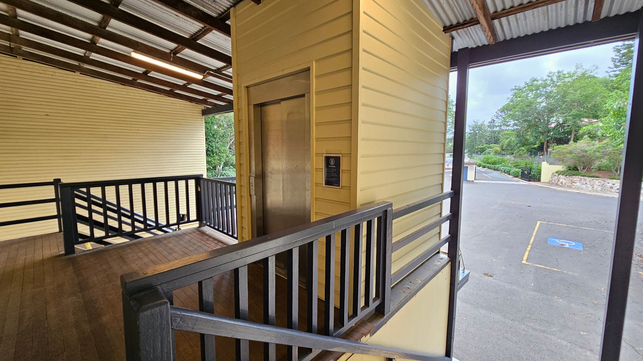 A wooden ramp with black railings leads to a lift with a metallic door on the left. The structure is painted yellow with horizontal panels. The ceiling is corrugated metal with exposed beams and a fluorescent light. On the right, there is an open area with a view of a paved road and a parking space marked with a wheelchair symbol. In the background, there are green trees and a stone wall.