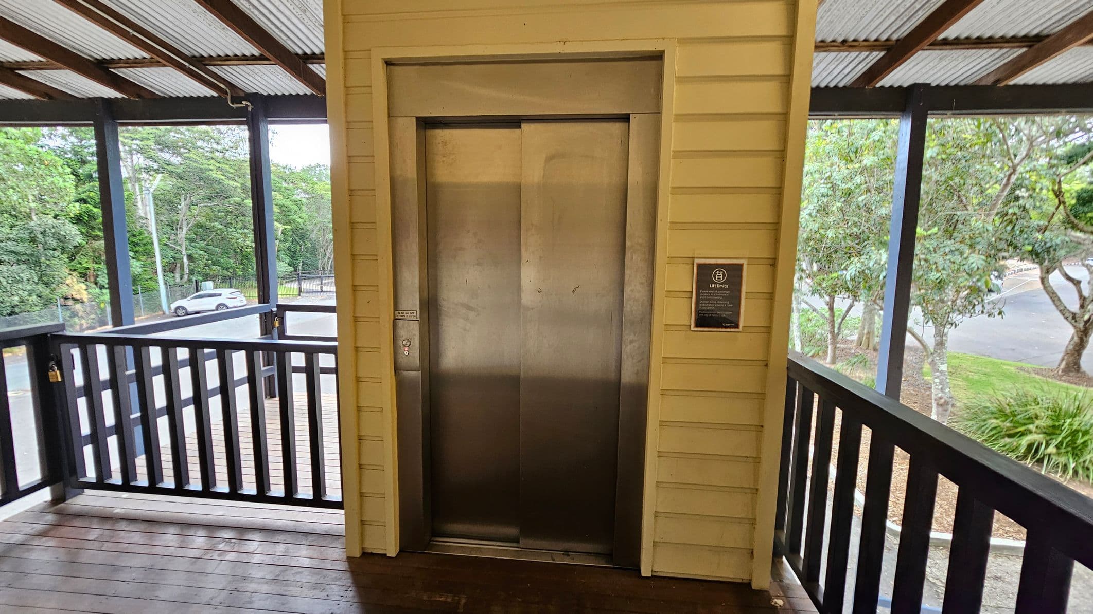 A metal lift door is centred on a wooden wall with a yellowish hue. To the right, a sign is mounted on the wall. The area is enclosed by a black railing, and beyond it, trees and a parked white car are visible. The floor is wooden, and the ceiling is made of corrugated metal with exposed beams.
