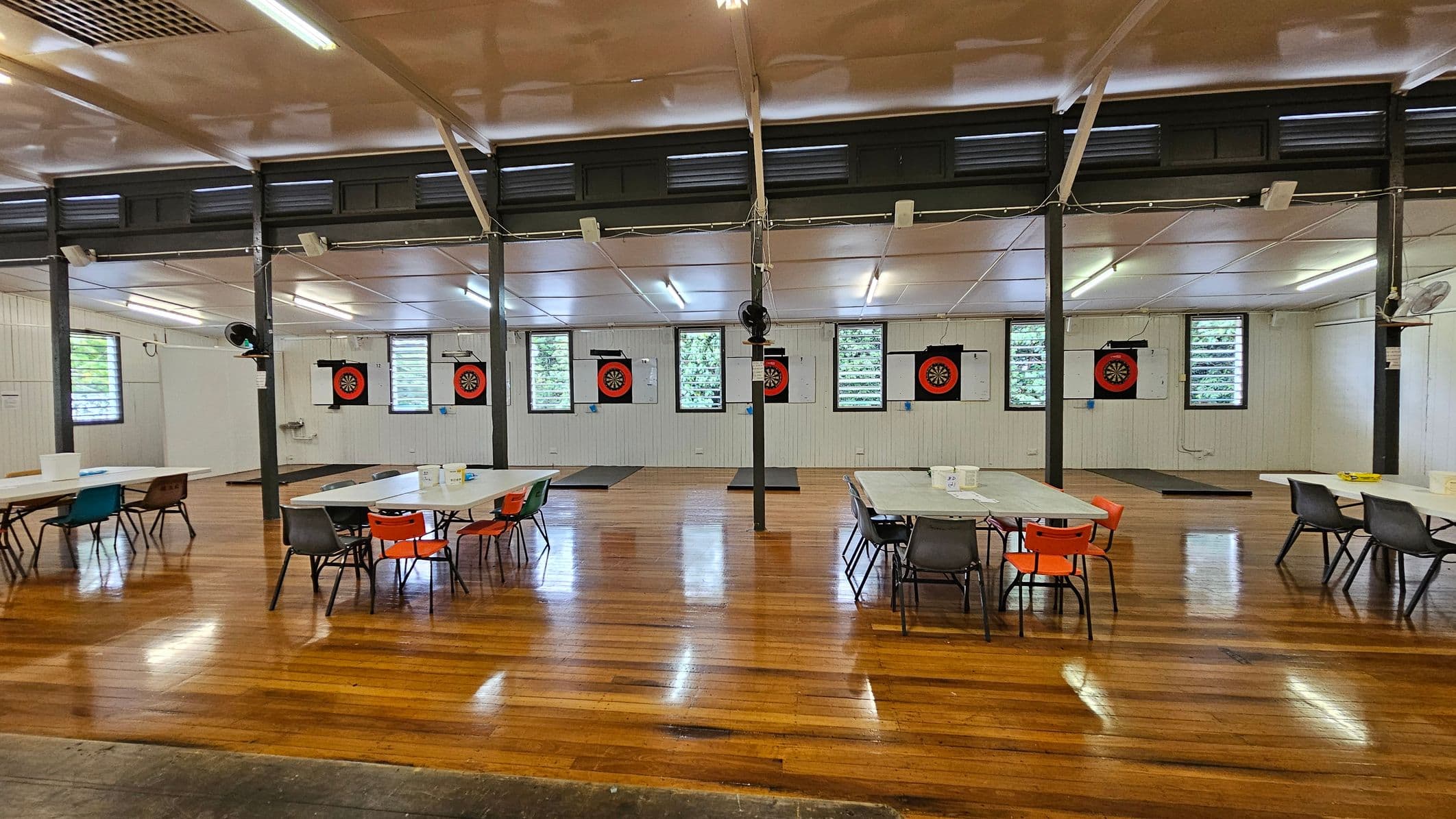A spacious hall with polished wooden floors and a high ceiling. On the left, there are dartboards mounted on the wall, with colourful pennants hanging above. In the centre, rows of tables and chairs are arranged neatly, featuring red and grey seating. The right side has windows with louvres, allowing natural light in. A wooden plaque is mounted on the far wall. The ceiling has fluorescent lights and ceiling fans.