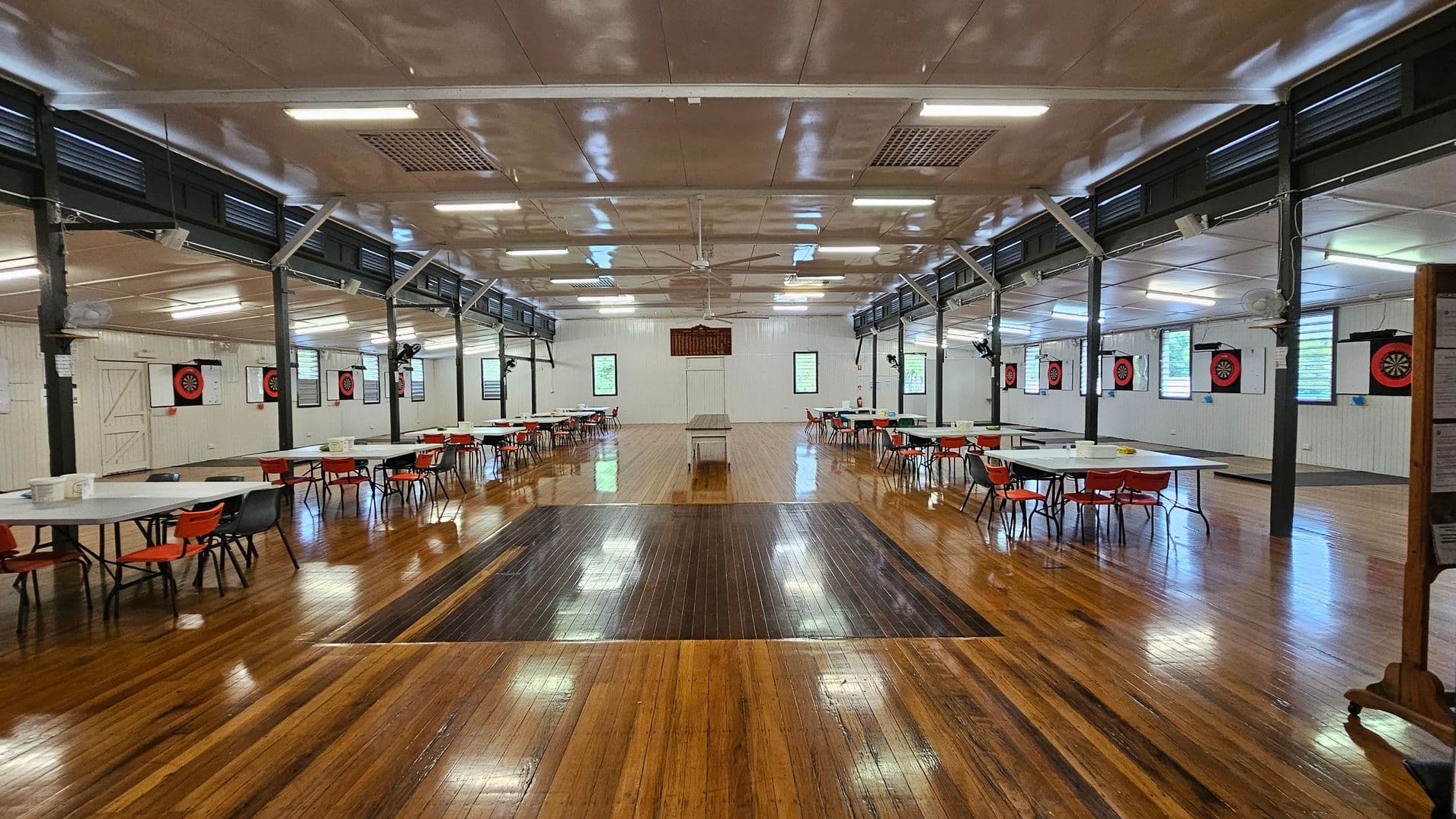 A spacious hall with a polished wooden floor and a white ceiling featuring fluorescent lights and ceiling fans. On the left, there are tables with red chairs arranged in rows. The right side has a notice board with various papers pinned to it. The back wall has dartboards and windows with green views outside. A long table is centred in the room.