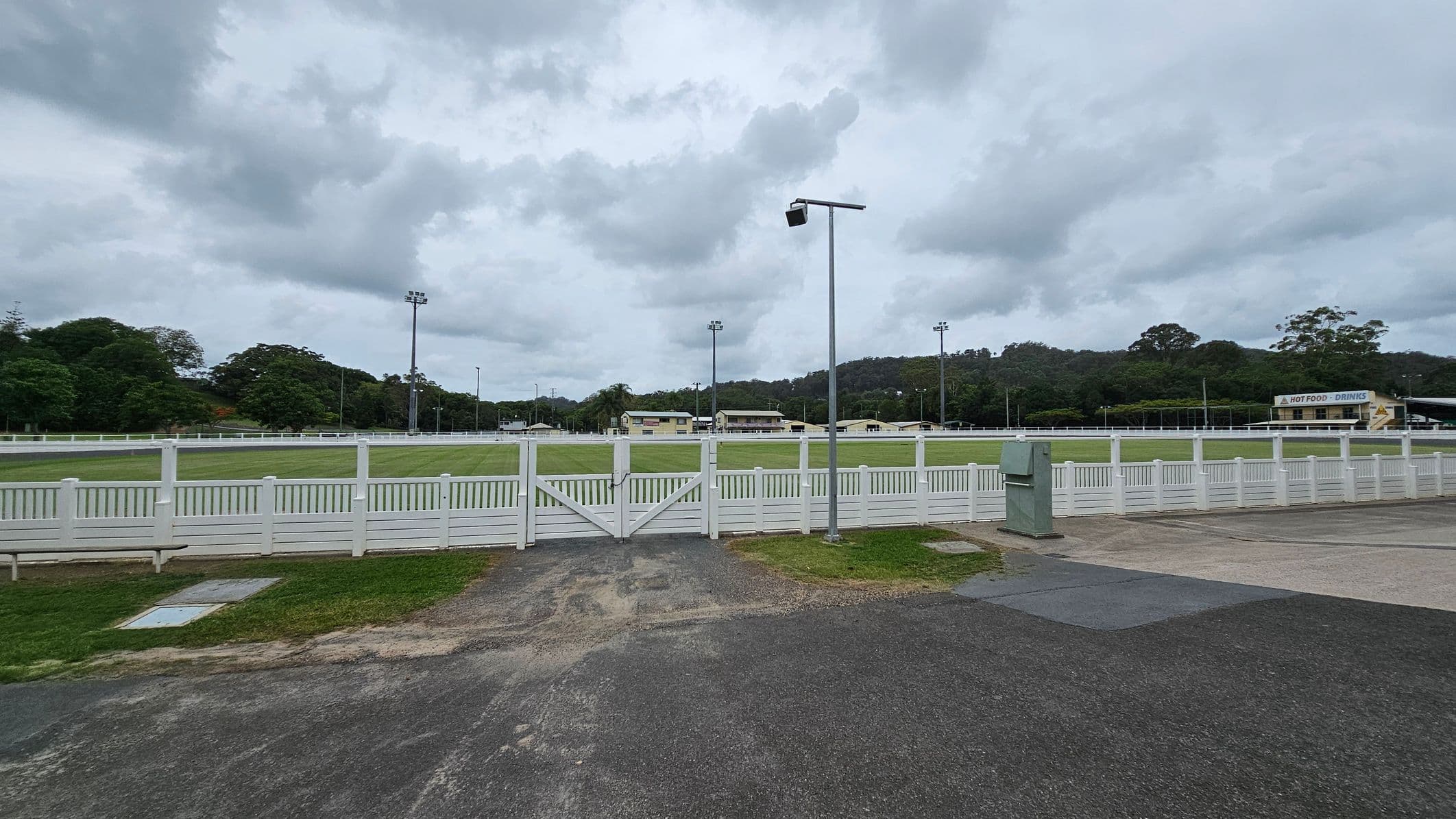 A sports field with a white fence runs across the middle, with a green lawn inside. Several floodlights are positioned around the field. In the background, there are buildings and trees on a hill. On the right, a yellow building with a sign for toilets is visible. The ground is paved with asphalt and concrete, and a bin is near the fence. The sky is overcast.