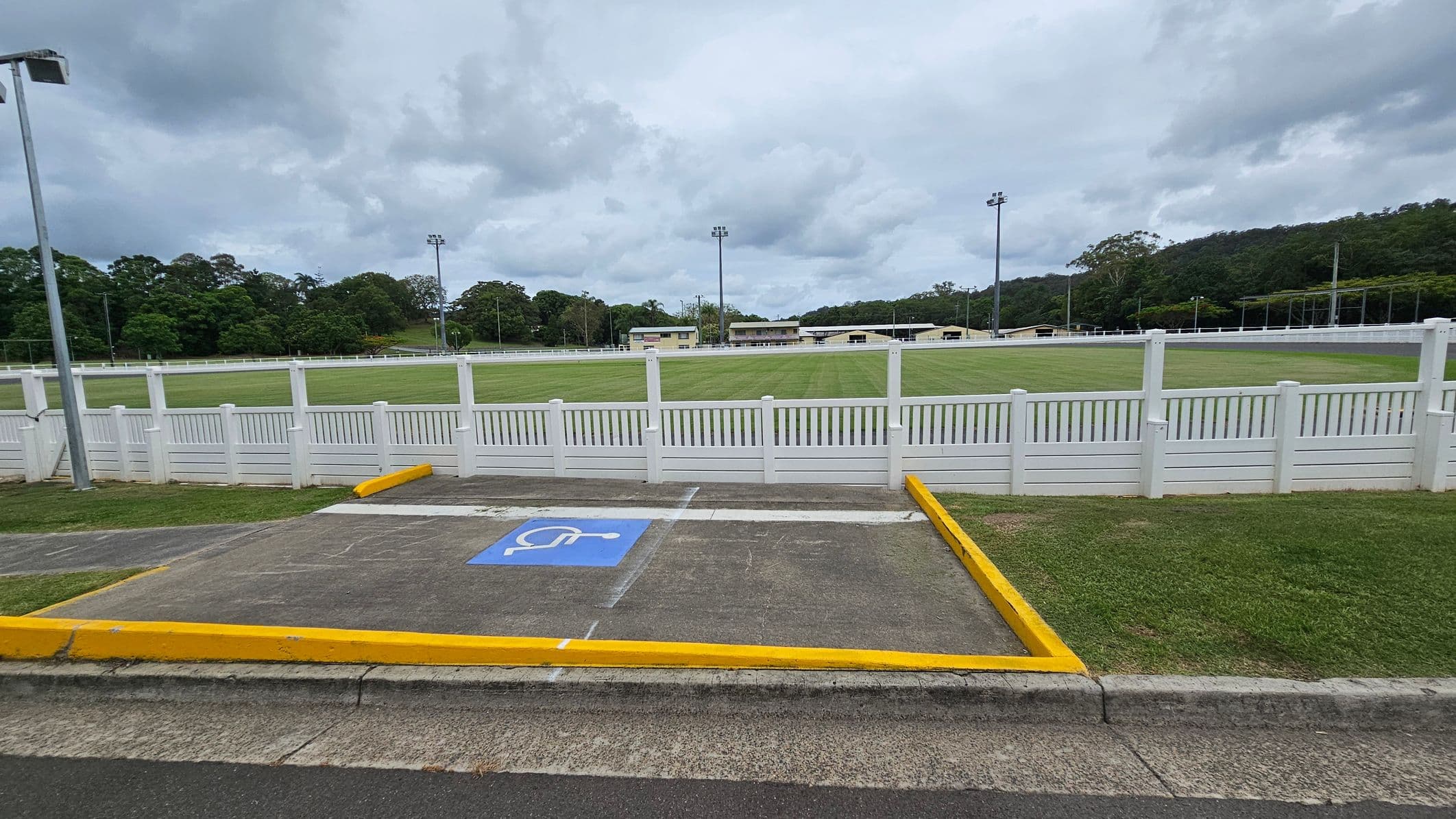 A grassy sports field is surrounded by a white picket fence. In the foreground, there is a parking space marked with a blue disabled symbol, bordered by yellow lines. The background features trees and several tall floodlights. A few small buildings are visible beyond the field. The sky is overcast.