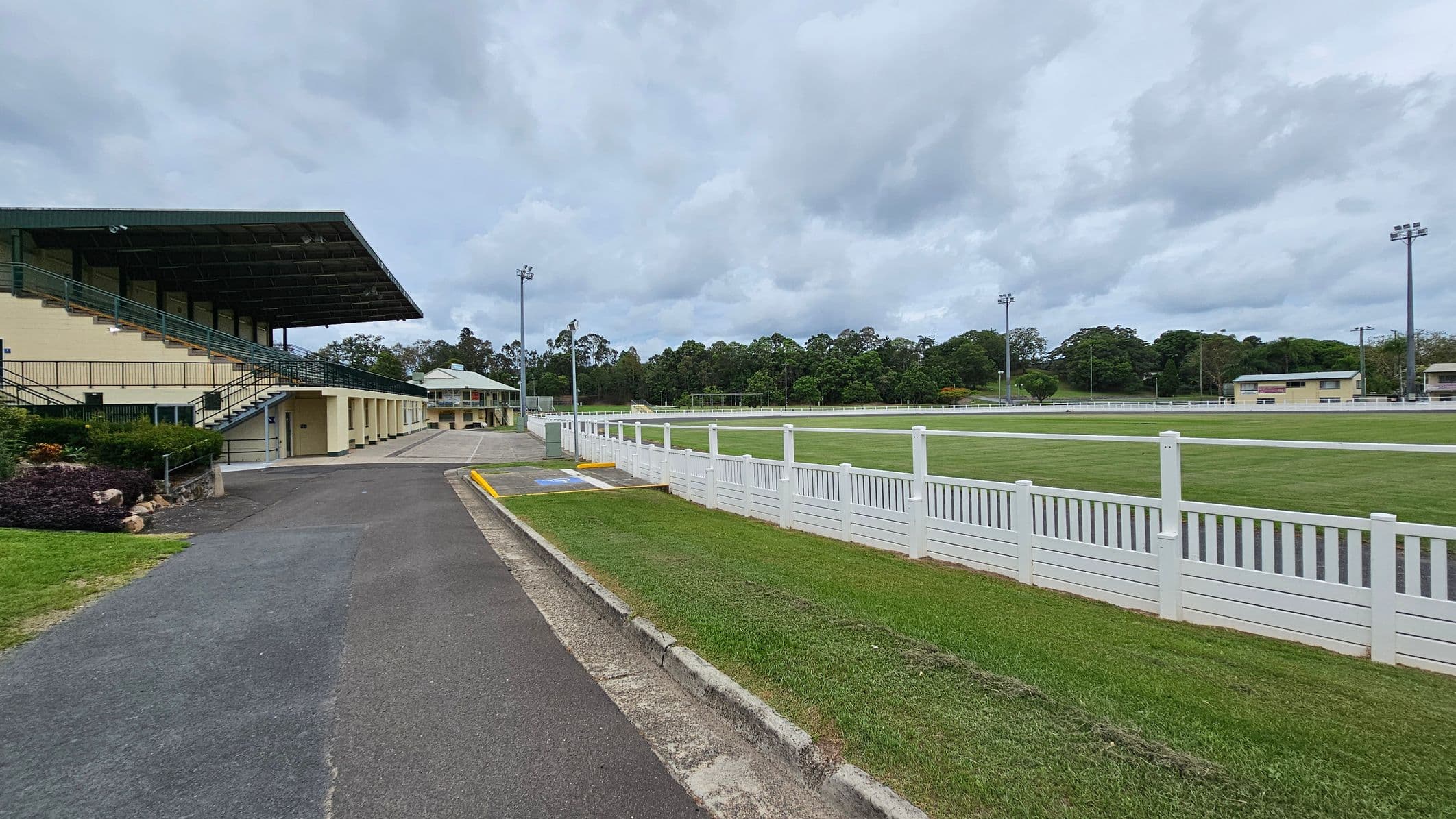 A sports field with a large grandstand on the left, featuring a green roof and multiple seating levels. A paved path runs alongside the grandstand, bordered by grass and a small garden. On the right, a white fence encloses a grassy field. In the background, there are trees and additional buildings, with tall floodlights visible around the field. The sky is overcast.
