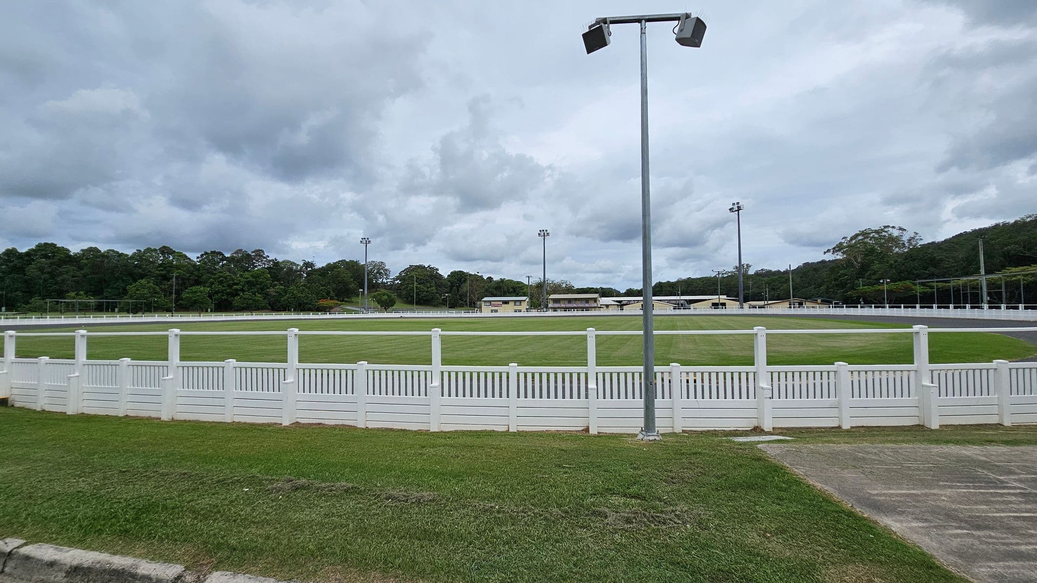 A grassy sports field is surrounded by a white picket fence. In the foreground, there is a patch of grass and a concrete path. A tall floodlight stands near the centre. In the background, there are several buildings and more floodlights. Dense trees line the horizon under a cloudy sky.
