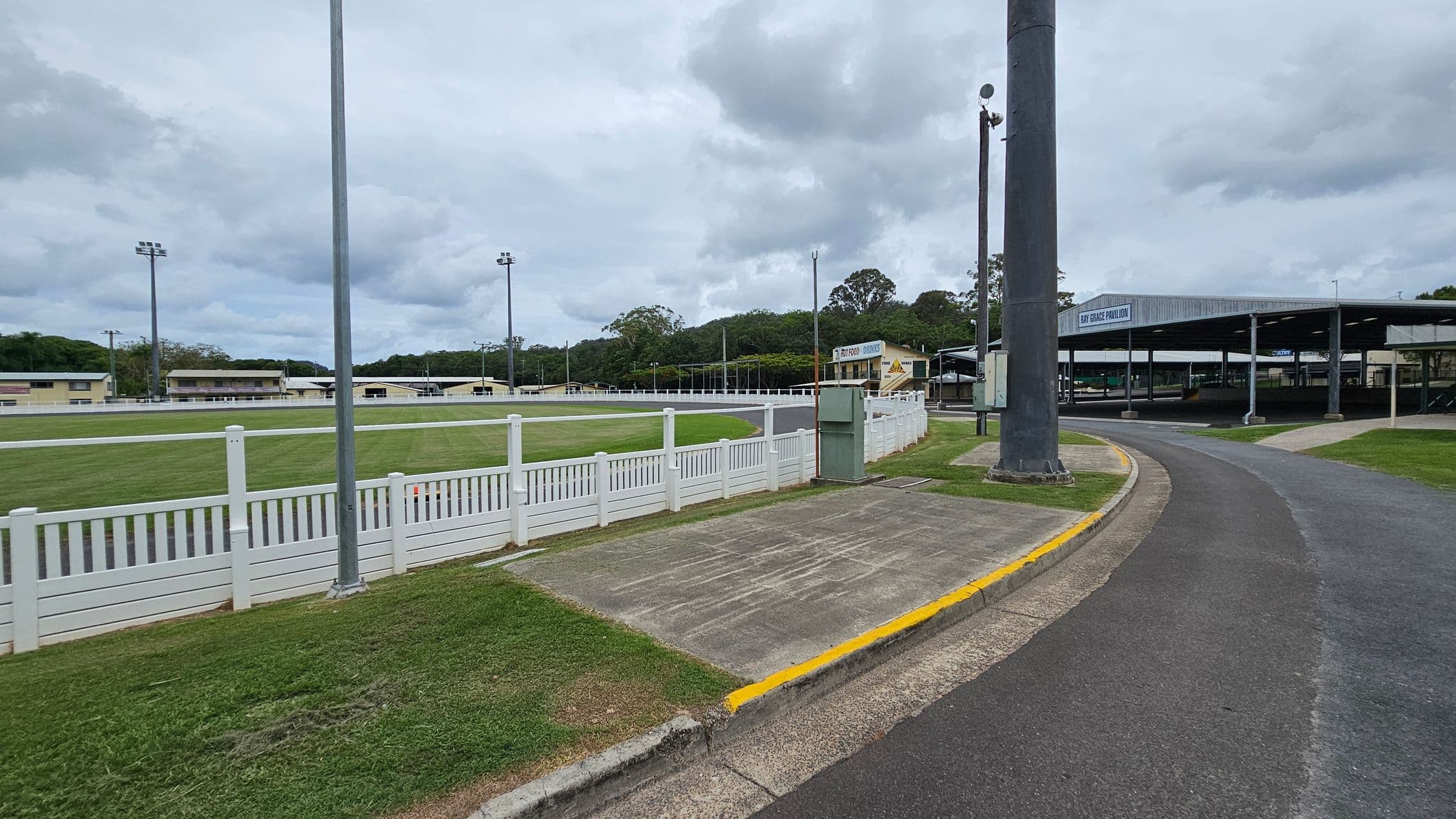 A paved road curves to the right, bordered by a grassy area and a white picket fence on the left. Beyond the fence is a green field with several tall floodlights. In the background, there are multiple low buildings, including one labelled "Bay Grace Pavilion." A large pole stands near the road, and trees are visible in the distance under a cloudy sky.