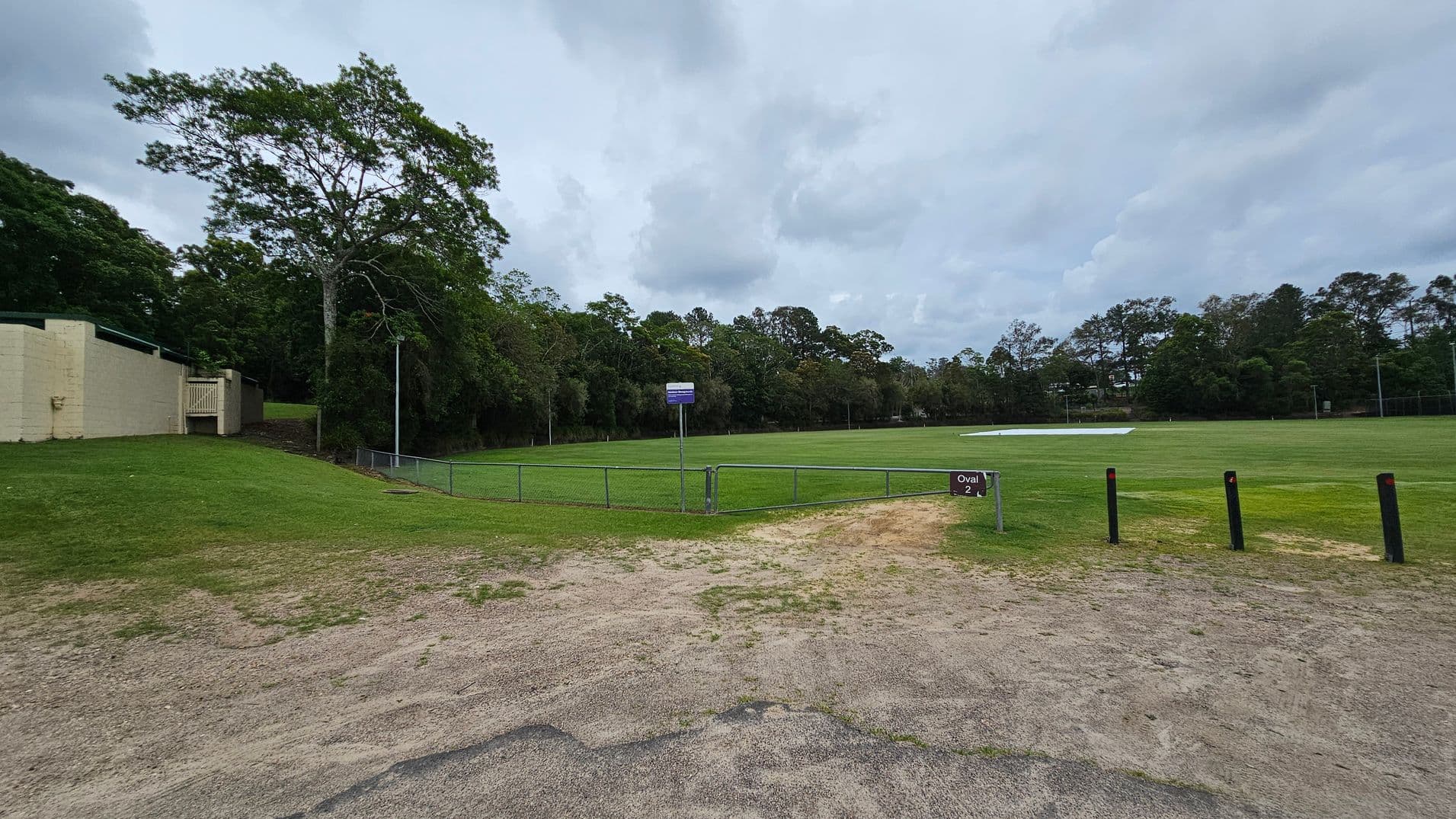 A grassy sports field with a sign reading "Oval 2" in the foreground. The field is surrounded by a low metal fence. On the left, there is a beige building with a small gate and a large tree nearby. The background features a line of dense trees. The ground in the foreground is a mix of grass and dirt, with several black bollards on the right side.