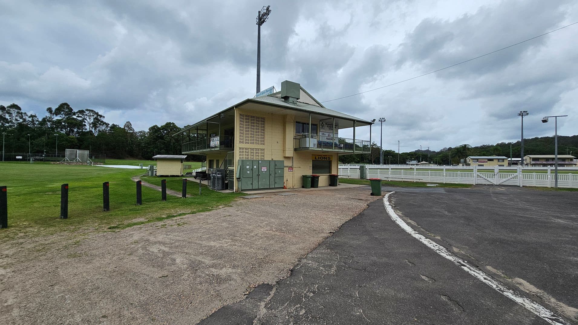 A sports field with a two-storey building on the right, featuring a sign that reads "LIONS" above a closed shutter. The building has a balcony with railings and several green bins nearby. The foreground has a mix of gravel and tarmac, leading to a curved road. On the left, there's a grassy field with black bollards and a netted practice area. In the background, there are trees and additional sports facilities, including floodlights and white fencing.