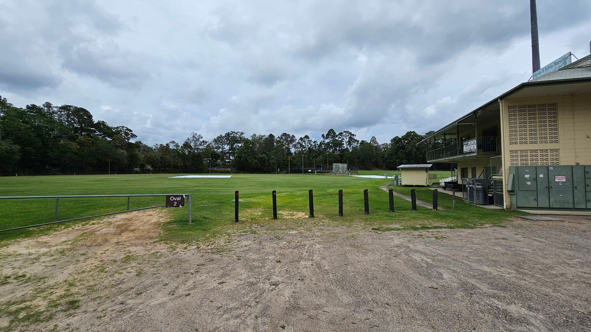 A grassy sports field labelled "Oval 2" is in the centre, surrounded by trees in the background. On the right, there is a two-storey building with a balcony and signs, alongside some utility boxes. The foreground has a mix of dirt and grass, with black bollards lining the edge of the field.