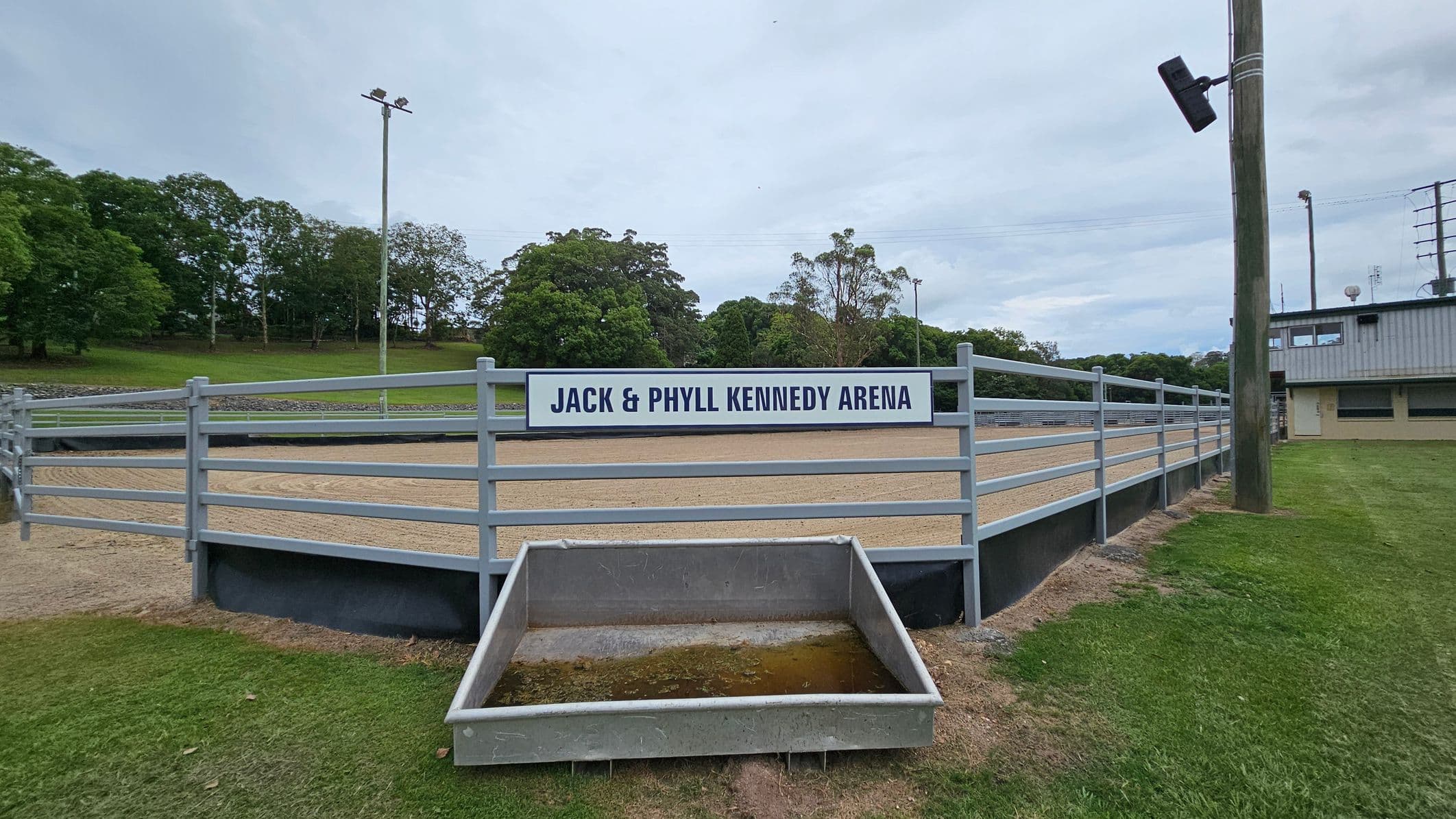 A fenced arena with a sign reading "Jack & Phyll Kennedy Arena" in the centre. The arena has a sandy surface and is surrounded by a metal railing. In the foreground, there is a rectangular concrete trough with some water. To the right, a wooden pole with a light fixture is visible, and a building with a corrugated metal roof is in the background. The left side features a grassy area with trees in the distance.