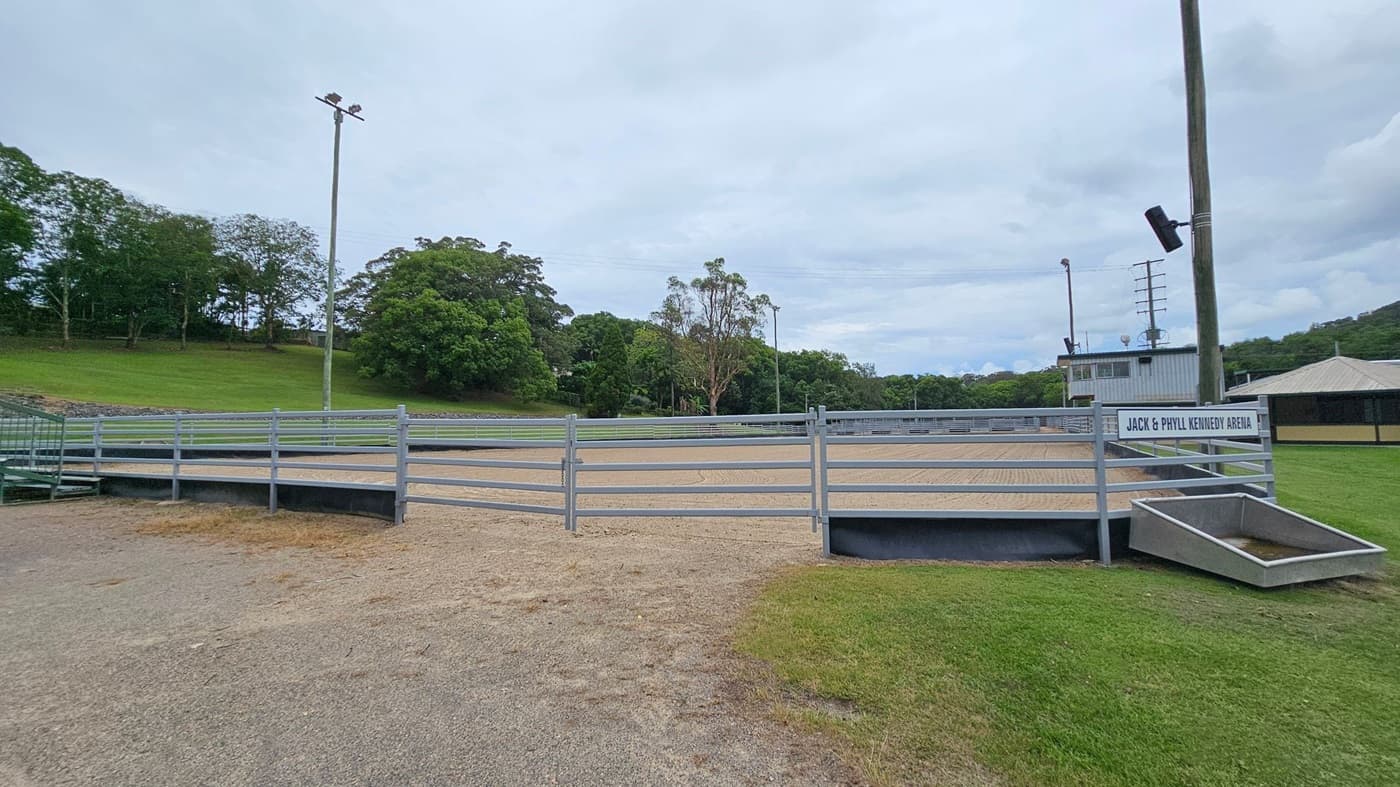 A large, sandy arena with visible rake patterns on the ground. On the left, there's a grassy embankment with dense green trees. On the right, a metal railing runs alongside a building with a corrugated metal roof and a sign partially visible. In the background, more trees and a few light poles are present. The sky is overcast.