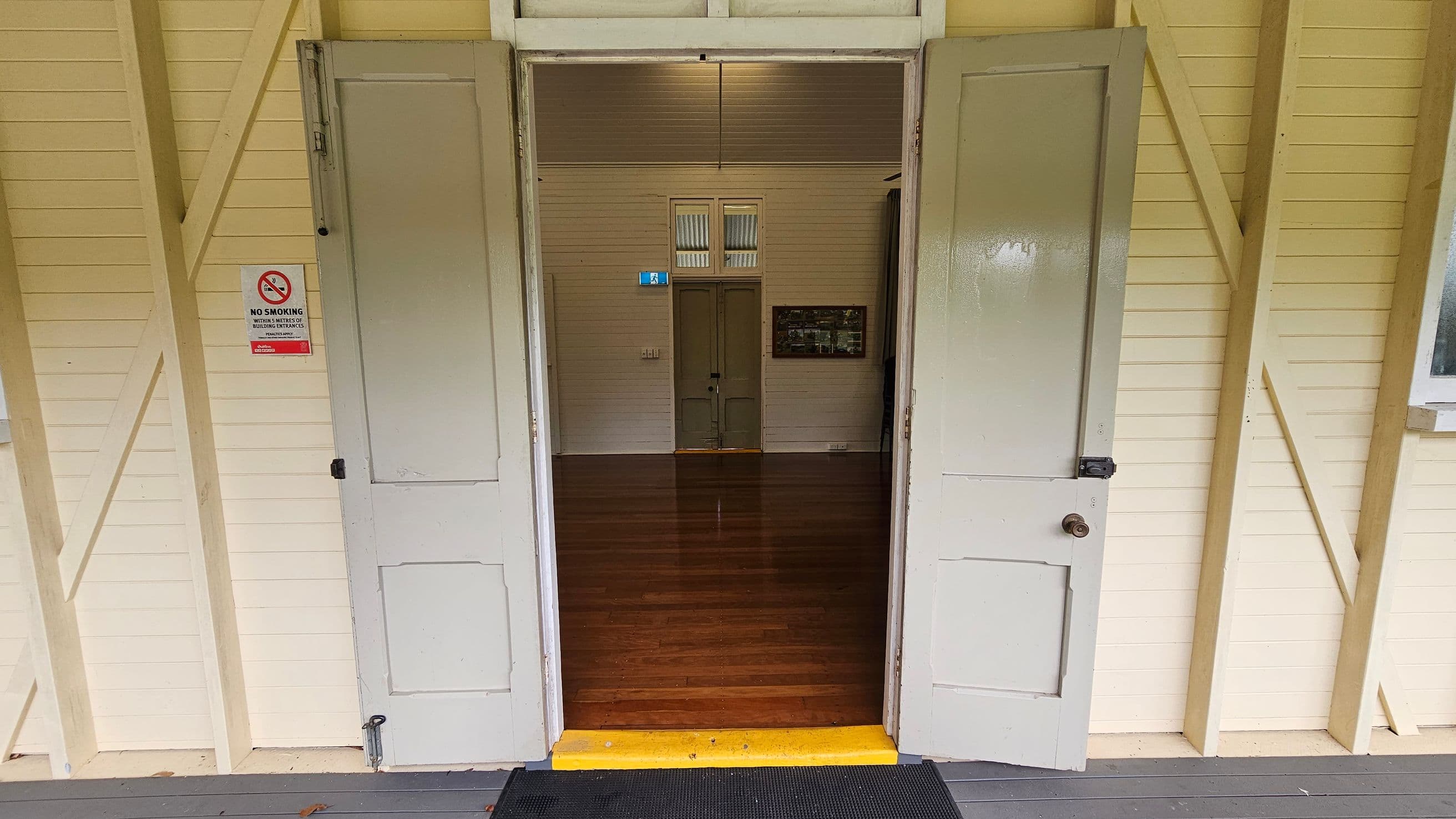 Open double doors lead into a room with polished wooden floors. The walls are cream-coloured with visible wooden beams. A "No Smoking" sign is on the left door. Inside, a closed door is visible at the far end, with a small window above it. A framed picture hangs on the right wall. The ceiling is wooden, matching the walls. A black mat and a yellow step are at the entrance.