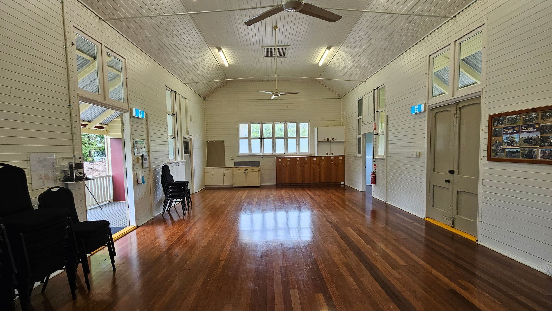 A spacious room with polished wooden floors and white panelled walls. On the left, there are stacked chairs and an open door leading to a porch. On the right, a closed door with a framed notice board nearby. The back wall features a row of windows above a series of cabinets, with a fire extinguisher to the side. The ceiling is high with two ceiling fans and fluorescent lights.