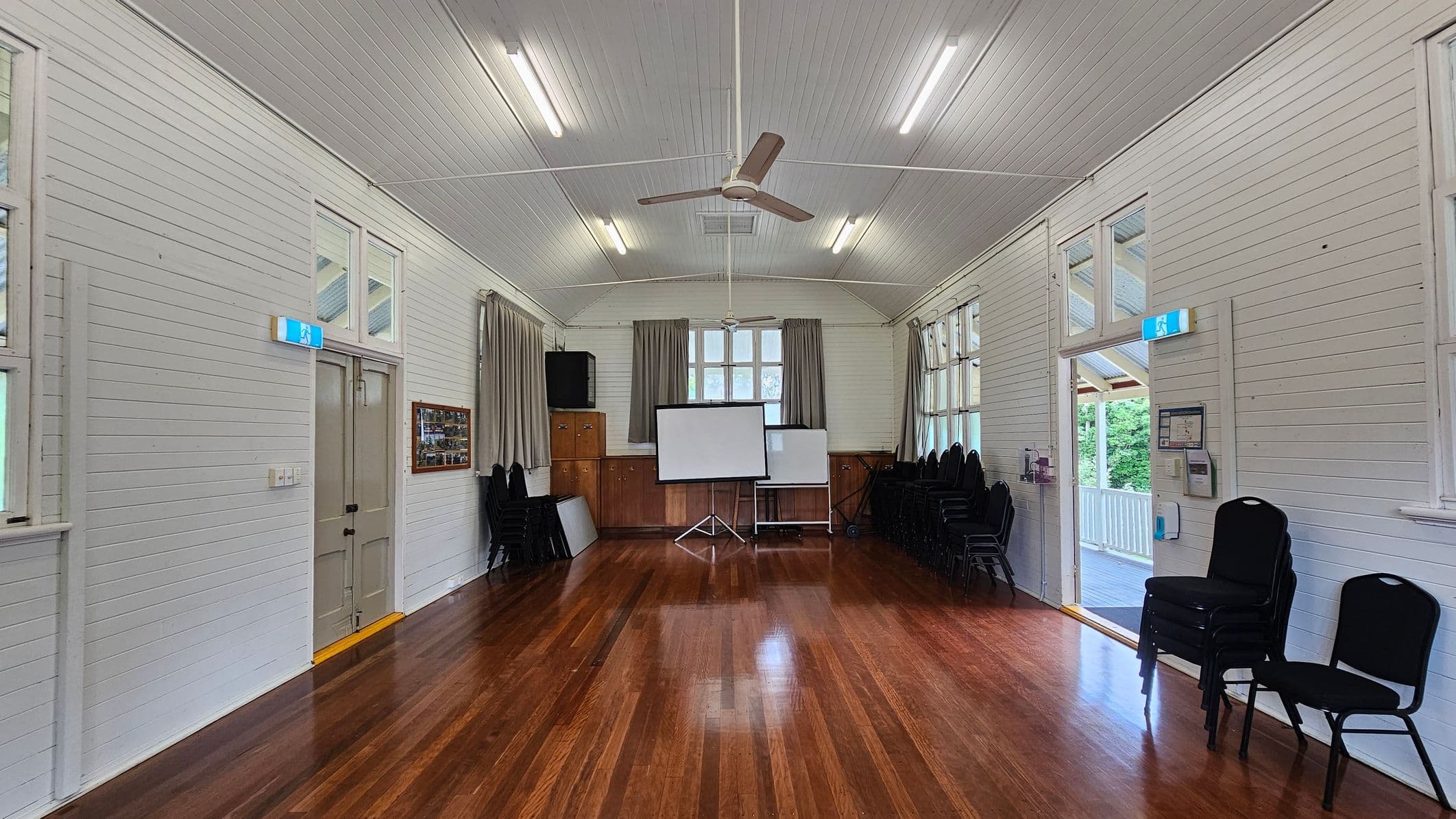 A hall with polished wooden floors and white panelled walls. In the centre, a projector screen stands in front of a window with curtains. On the left, a door with an exit sign above it, and a wall-mounted display. On the right, a stack of chairs and an open door leading to a balcony. The ceiling is vaulted with fluorescent lights and a ceiling fan.