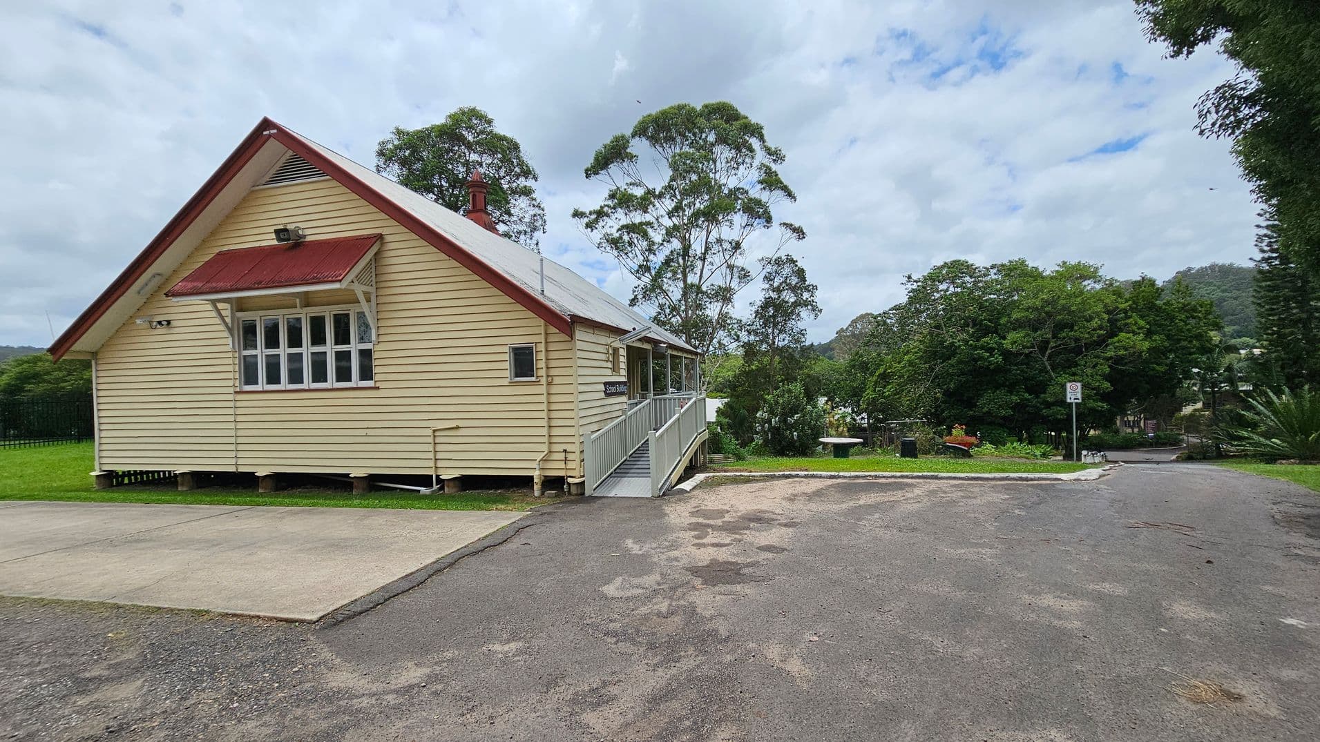 A small, single-storey wooden building with cream-coloured walls and a red roof is on the left. It has a window with an awning and a ramp leading to the entrance. The foreground features a paved area and a gravel path. In the background, there are tall trees and a grassy area with a bench and a signpost. The scene is surrounded by greenery, with hills visible in the distance.