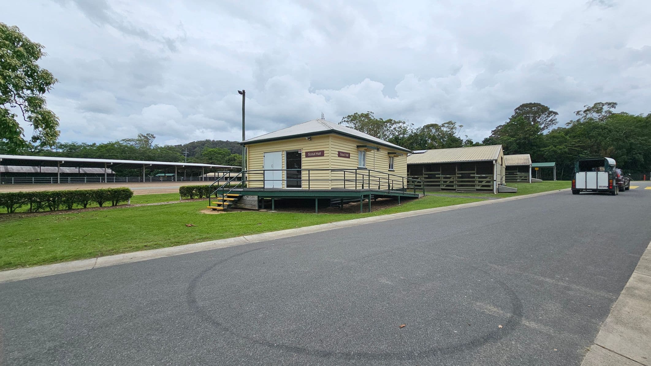 A small, elevated yellow building labelled "Scout Hall" is in the centre, with a ramp and steps leading up to it. To the right, a paved road runs alongside, with a vehicle towing a trailer parked further down. On the left, there are long, covered structures and a row of trimmed bushes. The background features dense green trees and a cloudy sky. The ground is a mix of grass and asphalt.