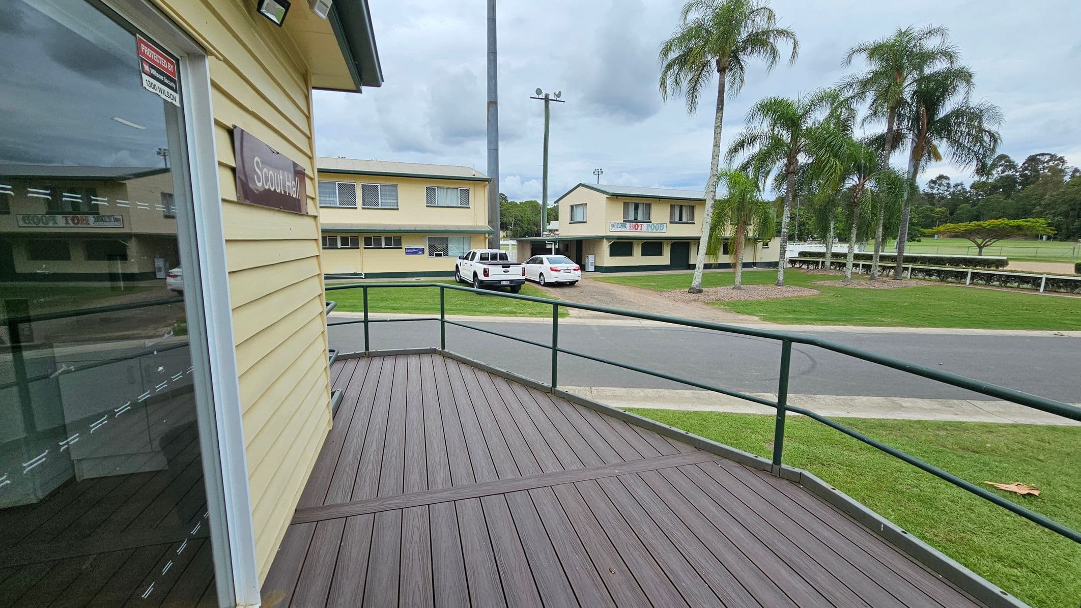 A view from a wooden deck with a green railing. On the left, a building with a sign reading "Scout Hall" is visible. In the background, there are two parked vehicles, a white pickup truck and a white car, in front of a yellow building with signs. To the right, a row of tall palm trees lines a grassy area. The scene includes a paved road and more greenery in the distance.