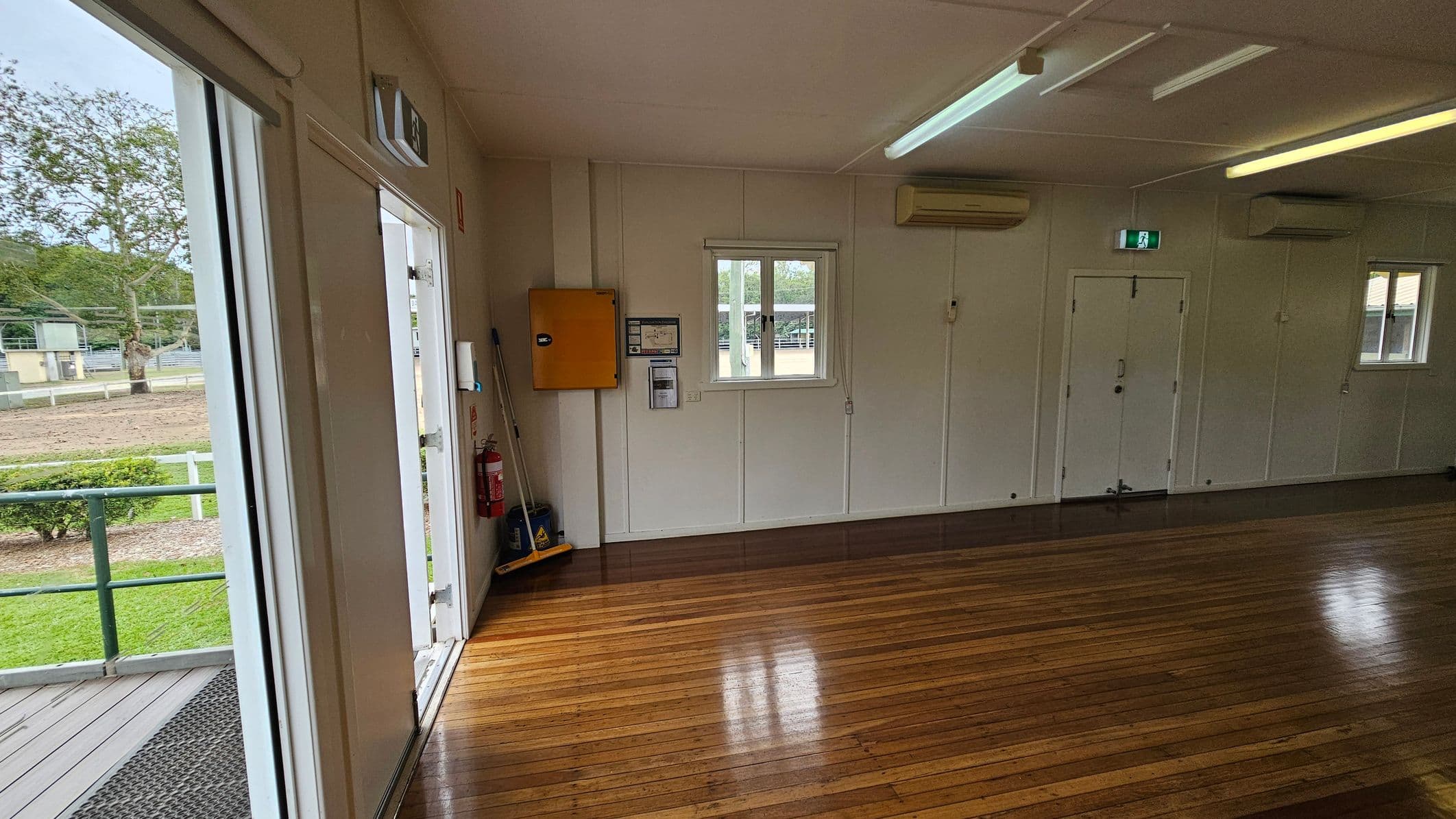 A room with polished wooden floors and white walls. On the left, there's an open door leading outside, showing a grassy area and a tree. Inside, a fire extinguisher and cleaning supplies are near the door. The back wall has a small window, a yellow cabinet, and an air conditioning unit. The ceiling has fluorescent lights, and there's an exit sign above a door on the right.