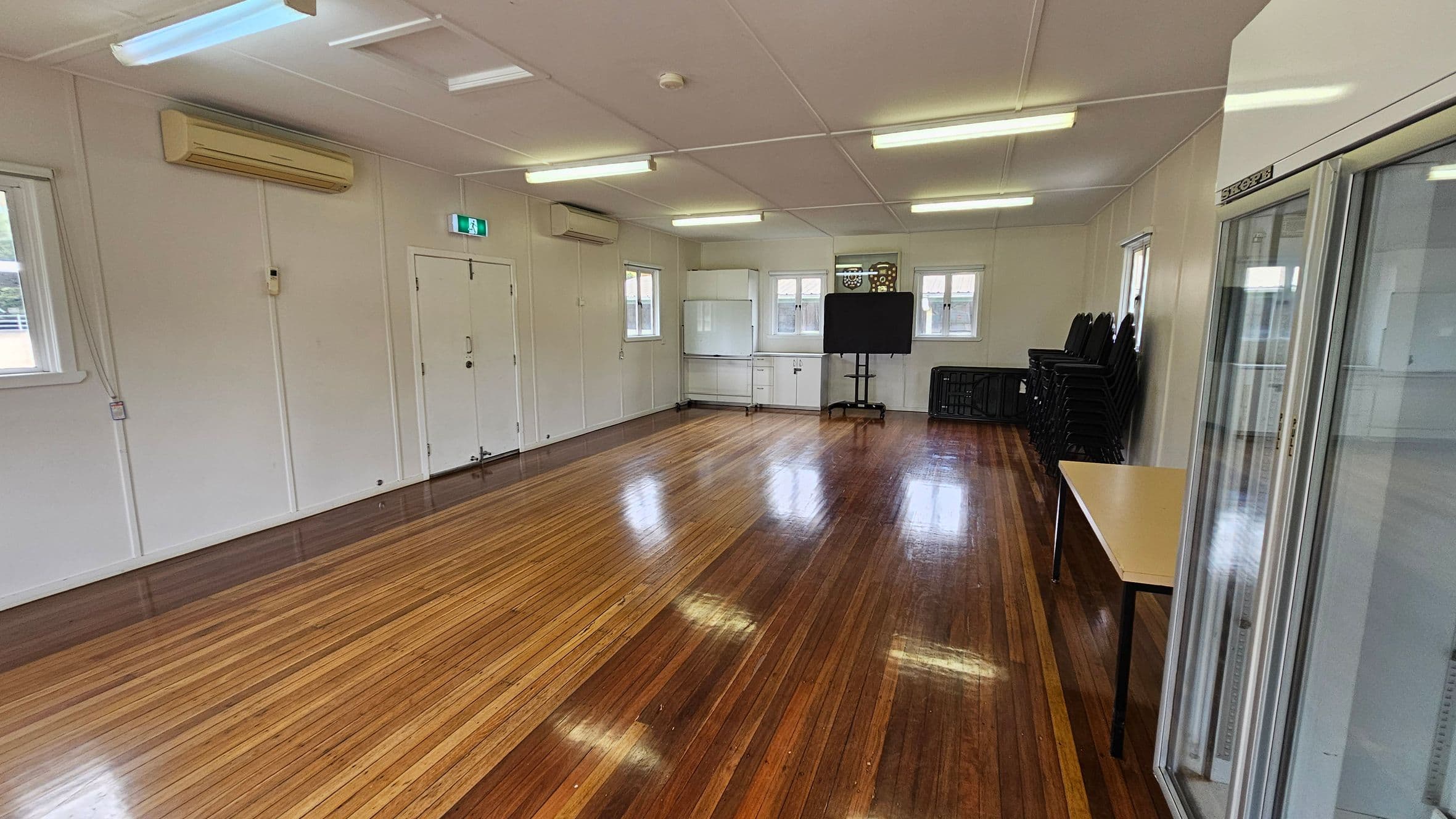 A spacious room with polished wooden floors and a white ceiling with fluorescent lights. On the left, there are white walls with air conditioning units and a double door. The back wall has windows, a white cabinet, and a black screen on a stand. On the right, there is a stack of black chairs, a folded table, and a glass-fronted refrigerator.