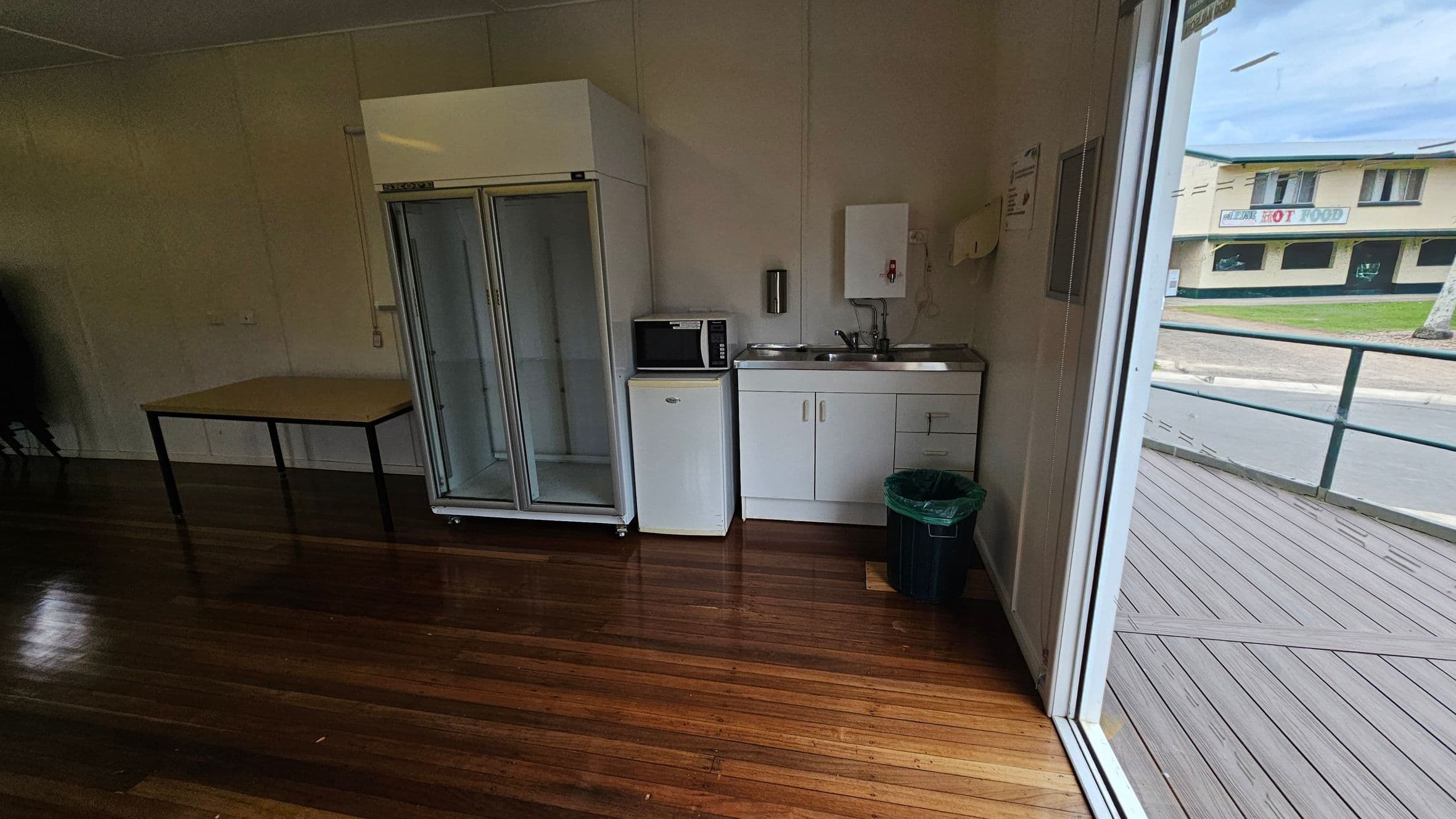 A small kitchenette with a wooden floor. On the left, there's a table and a large glass-fronted fridge. In the centre, a microwave sits atop a small fridge next to a sink with a cabinet below. A green bin is on the floor. On the right, a large window shows an outdoor deck and a view of a building across the street with a sign that reads "HOT FOOD".