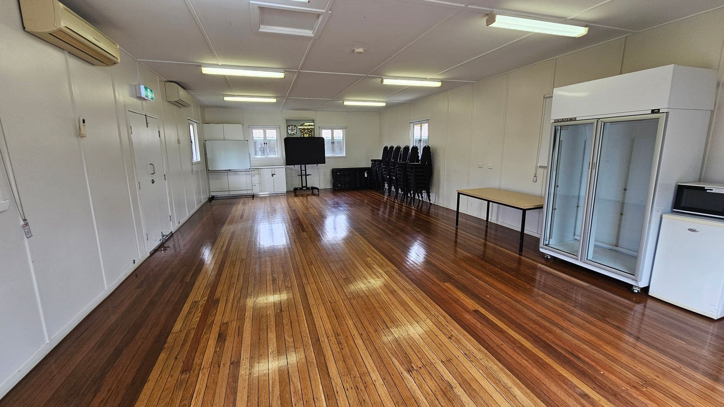 A spacious room with polished wooden floors and a white ceiling with fluorescent lights. On the left, there's an air conditioning unit and a door. In the middle, a blackboard and stacked chairs are against the wall. On the right, a large glass-fronted refrigerator, a table, and a small white fridge are visible. Windows are present on the far wall, allowing natural light in.