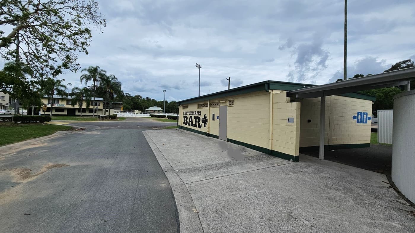A single-storey, cream-coloured building with green trims is on the right, displaying the text "Cattleman’s Bar" and directional signage. Adjacent to it, a paved concrete area transitions to a tarmac driveway. On the left, a tree with sparse branches is over a grassy area, and in the background, a multi-storey building with palm trees. The foreground features a slightly uneven tarmac surface, while tall light poles rise in the middle ground.