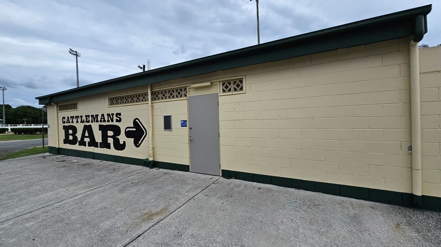 A cream-coloured single-storey building with green trims is on the right, featuring "Cattleman’s Bar" text and an arrow pointing right. A grey door and a ventilation block sit in the centre, with concrete paving on the foreground. The left shows part of a grassy area, a road, and some trimmed bushes. Tall light poles rise in the background. The ground is a concrete surface with scattered leaves, while the ceiling is dark green overhang roofing.