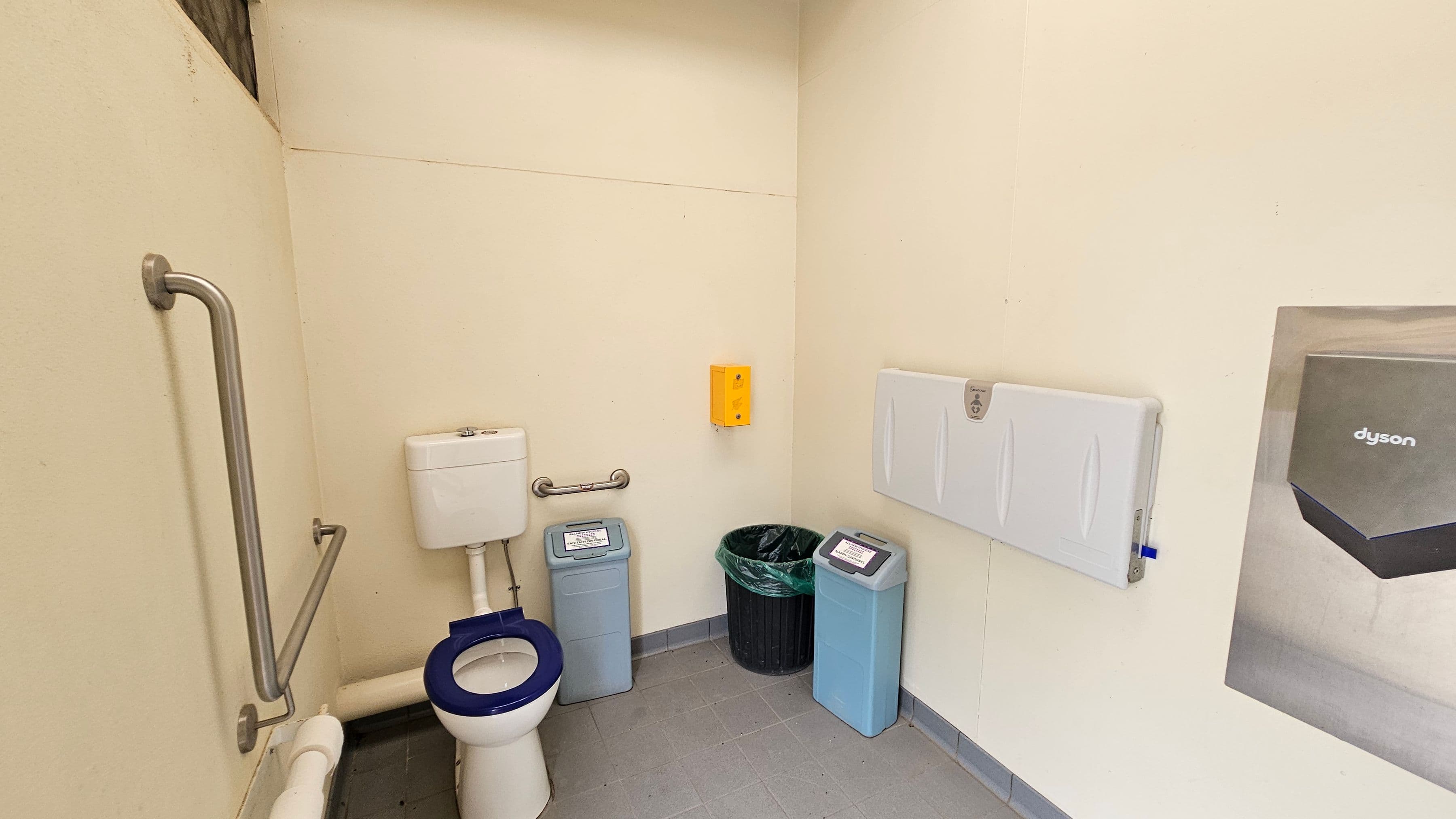 A white toilet with a blue seat is positioned on the left against cream walls, with stainless steel grab rails nearby. In the middle, two light blue bins and a black bin with a green liner are beneath a yellow dispenser. On the right, a folded white baby changing station is mounted on the wall beside part of a Dyson hand dryer. The floor is made of light grey tiles with dark grey grout, and part of the ceiling appears to be white with exposed beams in the upper left.