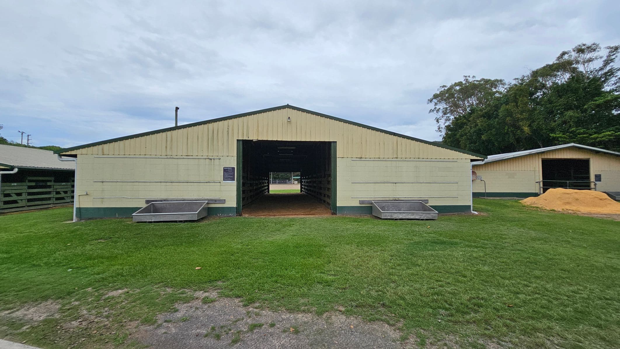 A light yellow barn with a green base stands in the centre, featuring a large open entrance. Two metal troughs are positioned on either side of the entrance. The ground is covered with grass, and a dirt path leads up to the barn. To the right, there's another similar structure with a pile of sand in front. Trees are visible in the background, adding greenery to the scene.