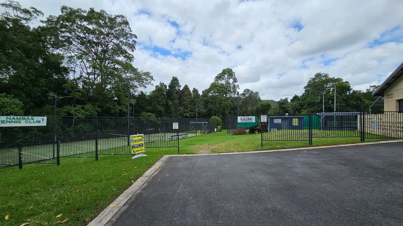A paved road curves on the right, leading to a building labelled "POULTRY LES DOWDLE PAVILION" with a light blue door. On the left, a tennis court is enclosed by a green fence, with trees in the background. A tree stands in the foreground on a grassy area, with scattered yellow leaves on the ground. A lamppost is next to the tree.