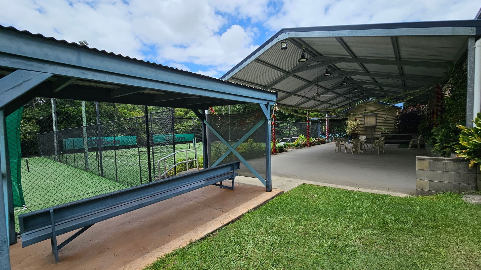 A covered outdoor area with a metal roof and open sides. On the left, there's a fenced tennis court with a green surface and a bench under a small shelter. On the right, a larger covered space with a concrete floor has several plastic chairs and tables. The background features lush greenery and a small wooden structure. The foreground has a patch of grass.
