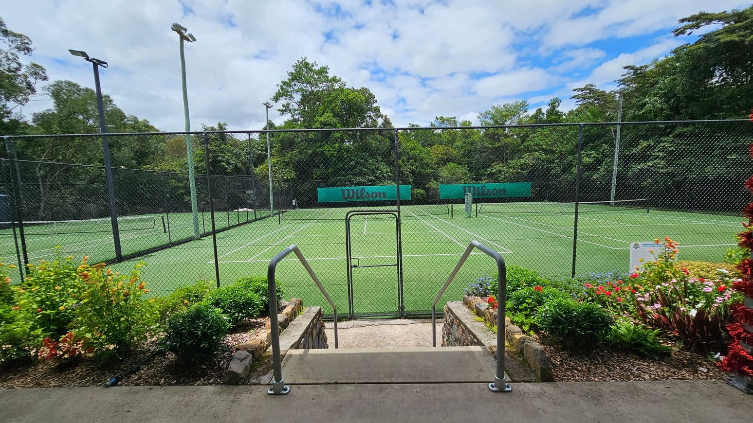 A tennis court with a green surface is surrounded by a chain-link fence. In the foreground, there are steps leading down to the court, flanked by metal railings. Colourful flowers and green shrubs line the sides of the steps. Tall floodlights are positioned around the court, and the background is filled with dense green trees. Two green banners with the word "Wilson" are visible on the far side of the court.