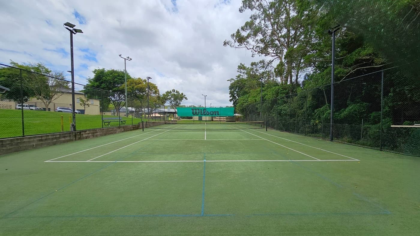 A tennis court with a green surface and white lines. In the centre, a net with a "Wilson" banner. On the left, a grassy area with trees and a building in the background. On the right, dense trees and foliage. Tall floodlights are positioned around the court, and a chain-link fence encloses the area.