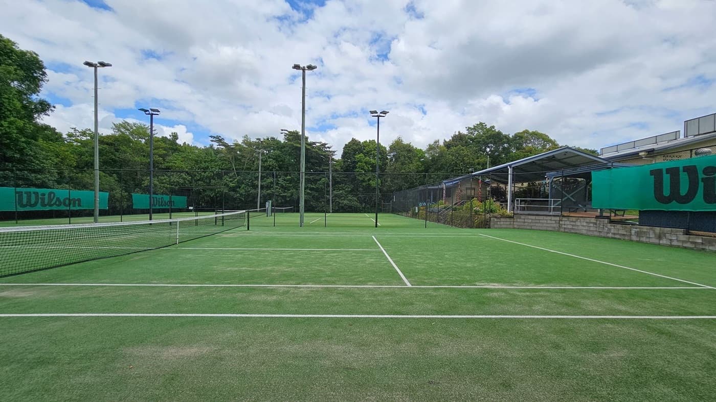 A tennis court with a green artificial grass surface and white lines. The net runs across the middle. On the left and right, green Wilson-branded banners are attached to the fences. Tall floodlights are positioned around the court. In the background, there are trees and a covered seating area with a metal roof and brick base on the right side.