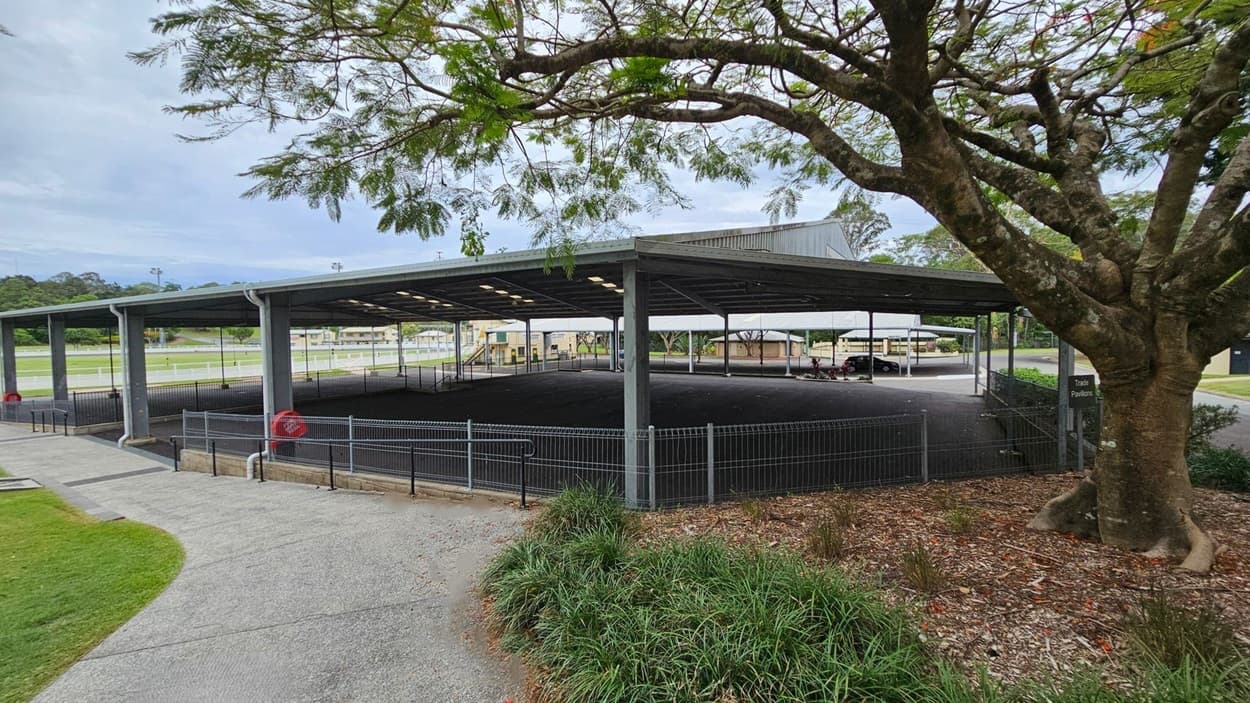 A large, open-sided pavilion with a metal roof and a sign reading "Ray Grace Pavilion" at the front. The structure is supported by metal beams and surrounded by a low hedge. In the background, there are grassy areas and additional buildings. A tree with green foliage is visible on the left. The ground is paved with asphalt, and there are signs and a small path leading to the pavilion.