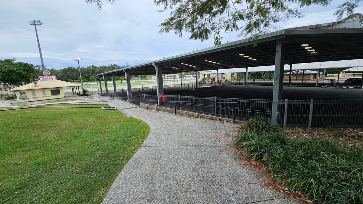 A paved path curves through a grassy area with a small building on the left displaying a "Hot Food Drinks" sign. To the right, a large covered structure with a metal roof and open sides is visible, surrounded by a fence. In the background, there are trees and a sports field. A tall light pole stands near the building, and some foliage is visible at the top edge of the image.
