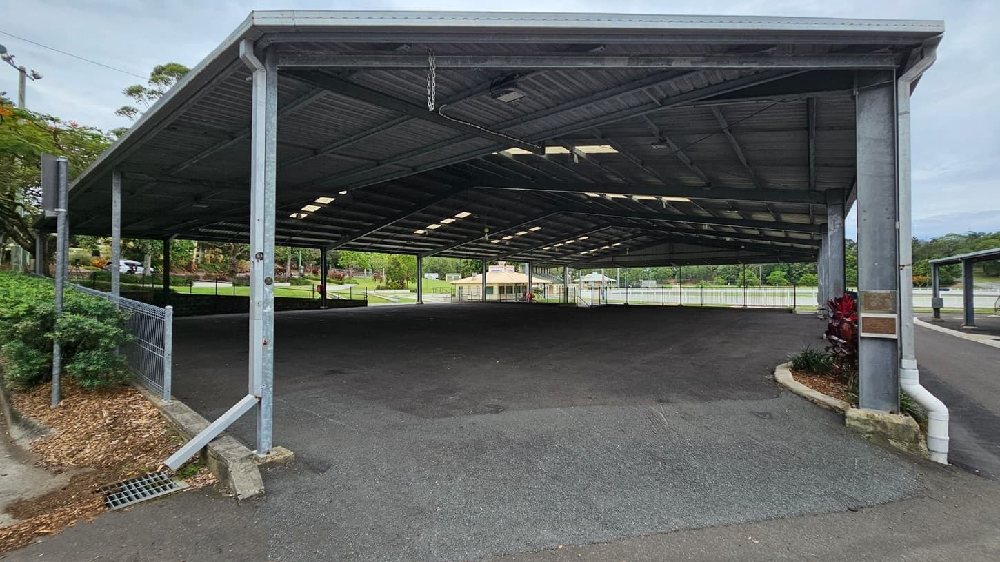 A large, open-sided metal shelter with a corrugated roof supported by metal beams. The ground is paved with asphalt. On the left, there is a fence with greenery behind it. In the background, a small building is visible, surrounded by trees and a grassy area. On the right, a small garden bed with red and green plants is next to a drainage pipe. The ceiling has several skylights allowing natural light to enter.