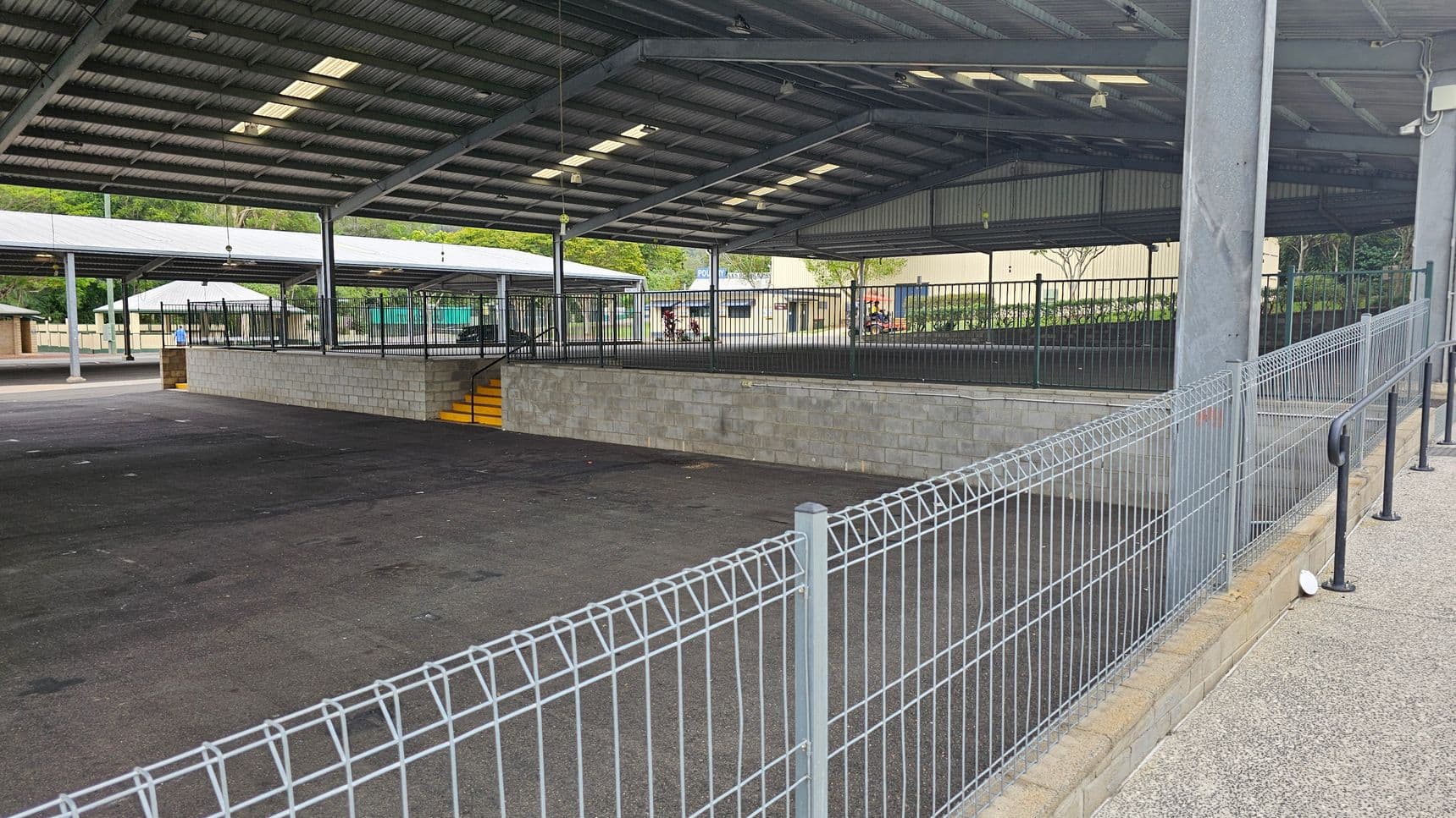 A large, open-sided structure with a metal roof and several support beams. The floor is paved with dark asphalt. In the centre, there is a raised platform with a low brick wall and yellow steps leading up to it. Metal fencing surrounds the platform. In the background, there are trees and a building with a sign partially visible. A wire fence runs along the right side, with a handrail attached to a low wall.