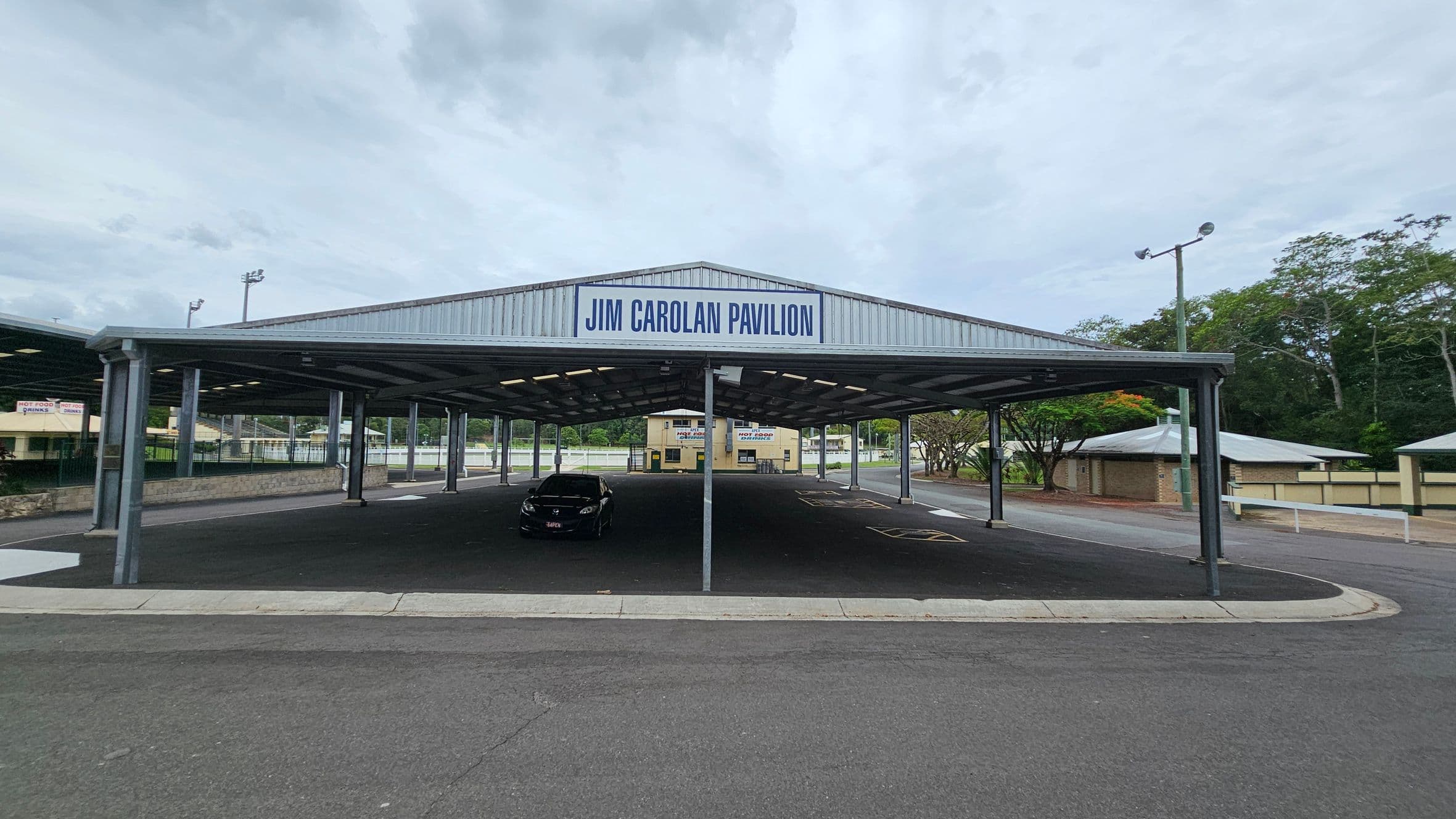 A large, open-sided pavilion with a metal roof labelled "Jim Carolan Pavilion" at the top. Underneath, a single black car is parked on the dark asphalt surface. In the background, there are trees and a few small buildings, including a yellow one with signs. The area is surrounded by a low fence and there are streetlights on tall poles. The ground is paved, and the pavilion is supported by metal columns.
