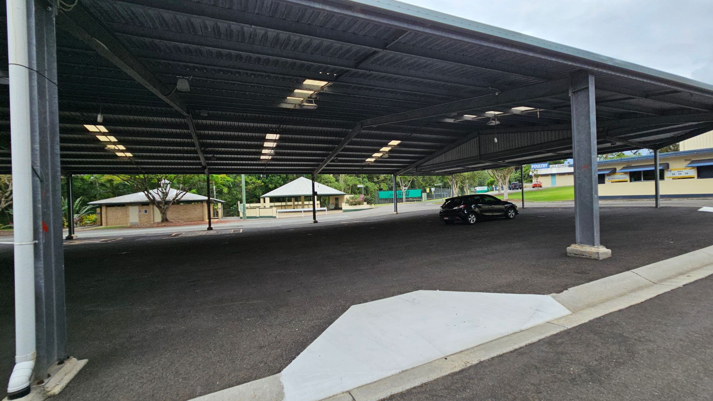 A covered parking area with a dark asphalt floor and a metal roof supported by columns. A single black car is parked on the right side. In the background, there are small buildings with light-coloured walls and green roofs, surrounded by trees and greenery. The ceiling has rows of fluorescent lights.