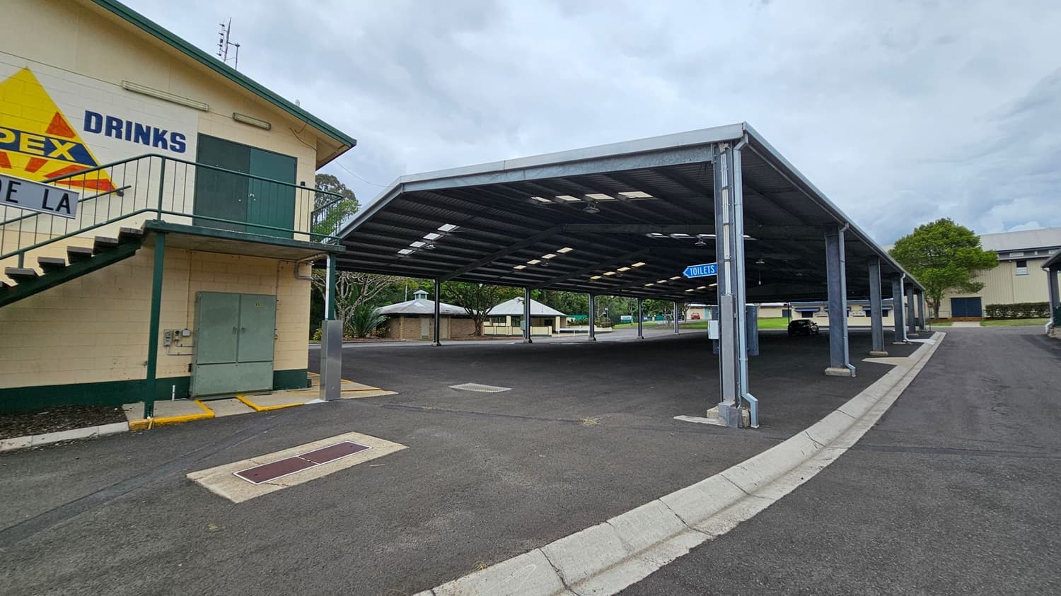 A paved area with a covered structure supported by metal beams on the right. A sign indicating "TOILETS" hangs from the structure. On the left, a building with a staircase and a sign that reads "DRINKS" is visible. In the background, there are trees and small buildings with green roofs. The ground is asphalt, and the area appears to be part of a larger complex or park.