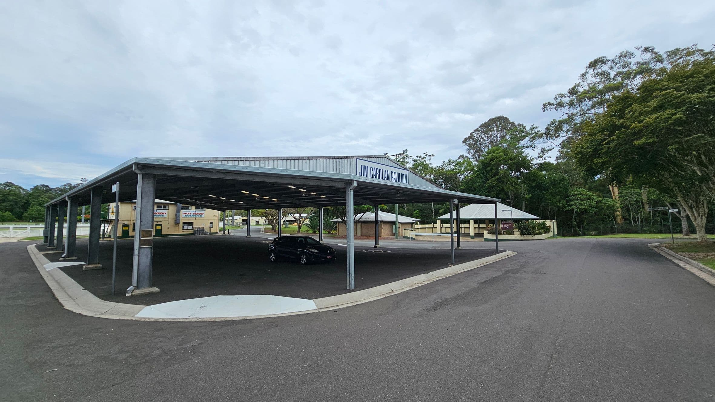 A large open pavilion with a metal roof labelled "Jim Carolan Pavilion" is on the left. Underneath, a black car is parked. To the right, a paved road curves around the structure, bordered by trees and greenery. In the background, there are small buildings, including a food stand with signs for "Hot Food" and "Cold Drinks". The area is surrounded by trees and vegetation.