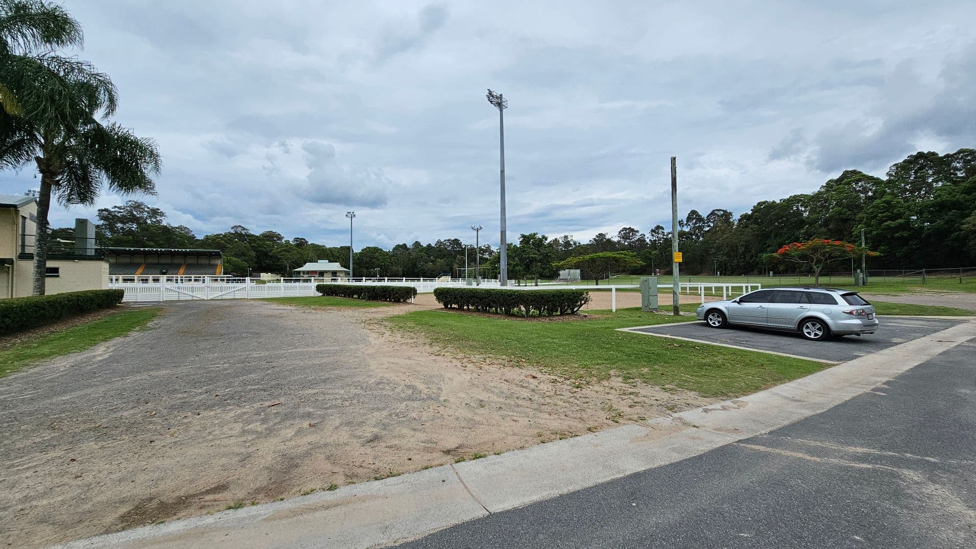 A sports field with a large light pole in the centre. On the left, there's a building with a palm tree nearby. A white fence surrounds the field, and there are hedges along the path. On the right, a silver car is parked in a designated parking area. The background features trees and additional light poles. The ground is a mix of grass and dirt, with a paved road in the foreground.