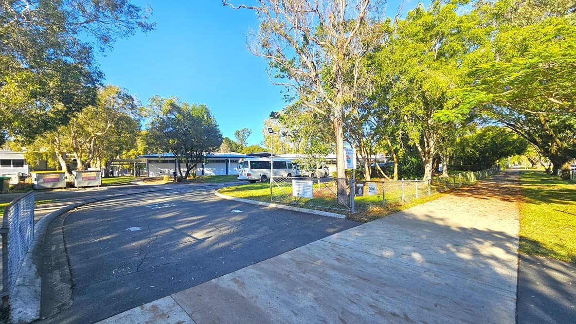 On the left, there is a large building with a sign reading "NAMBOUR SPECIAL SCHOOL" in bold blue letters. The building is partially covered by a tall green hedge. On the right, there is a white sign with the same logo and text, next to a tree-lined path. In the foreground, there is a concrete pavement and a metal fence running parallel to the hedge. The background features more trees and greenery. The floor is a mix of concrete pavement and grass.