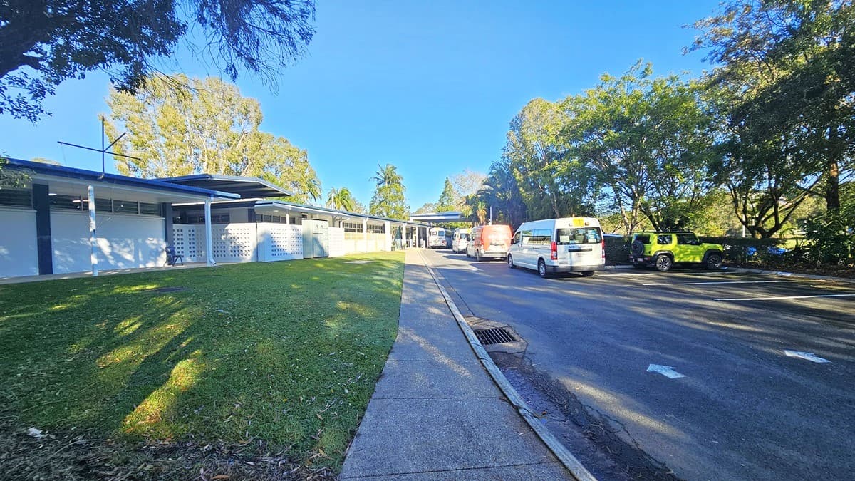 On the left, there is a grassy slope leading up to a long, single-storey building with a white facade and blue trim. On the right, there is a paved road with several parked vehicles, including a white van and a green car. In the middle, the road and footpath continue towards the background, where more trees and parts of the building are visible. The floor consists of grass on the left and asphalt on the right.