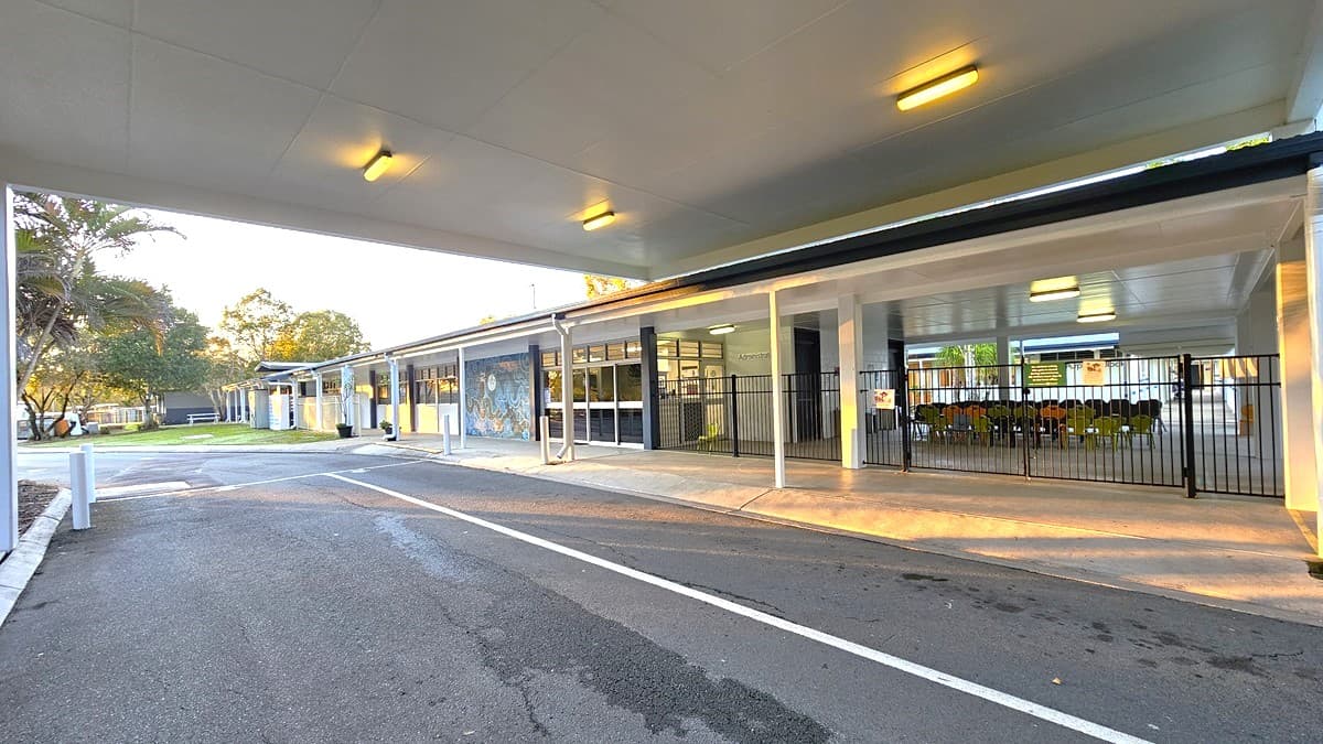 On the left, there is a long building with large windows and a mural near the entrance. On the right, there is a fenced area with a gate, leading to a covered walkway with tables and chairs inside. In the middle, the covered area extends towards the background, showing more of the building and trees. The floor is a mix of asphalt and concrete pavement.