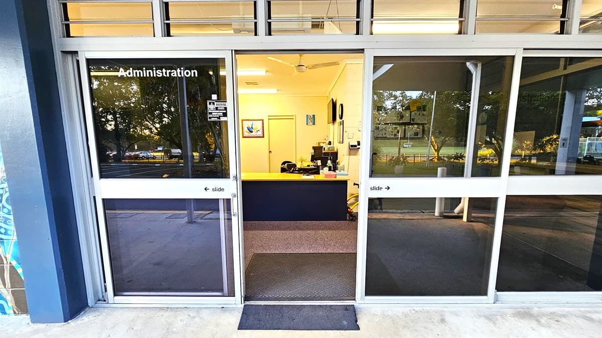 On the left, there is a blue wall with part of a mural visible. On the right, there is a large glass window reflecting trees and a parking area. In the middle, there are glass double doors labelled "Administration," leading to an office with a desk, computer, and wall art. The floor outside is concrete, transitioning to carpet inside.