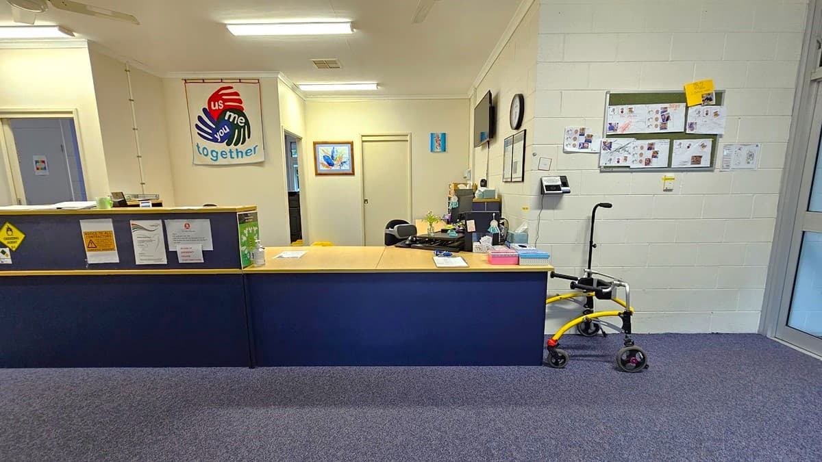 On the left, there is a blue reception desk with various notices and a green plant on top. Behind the desk, there is a wall with a colourful "Together" sign featuring handprints. On the right, there is a white wall with bulletin boards displaying papers and a yellow walker parked against it. In the middle, there is a doorway leading to an office with a desk and computer. The floor is covered in blue carpet, and the ceiling is white with fluorescent lights and a ceiling fan.
