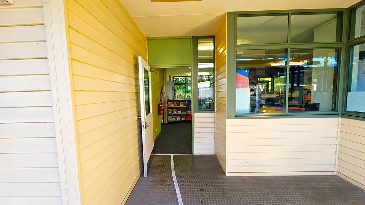 On the left, there is a yellow wooden wall with a white door that is slightly open. In the middle, the door leads to an interior space with bookshelves and a carpeted floor. On the right, there is a large window with green frames, providing a view of the interior. The background includes more bookshelves and a glimpse of colourful items. The floor outside the door is grey and textured, while the ceiling is white with a light fixture.